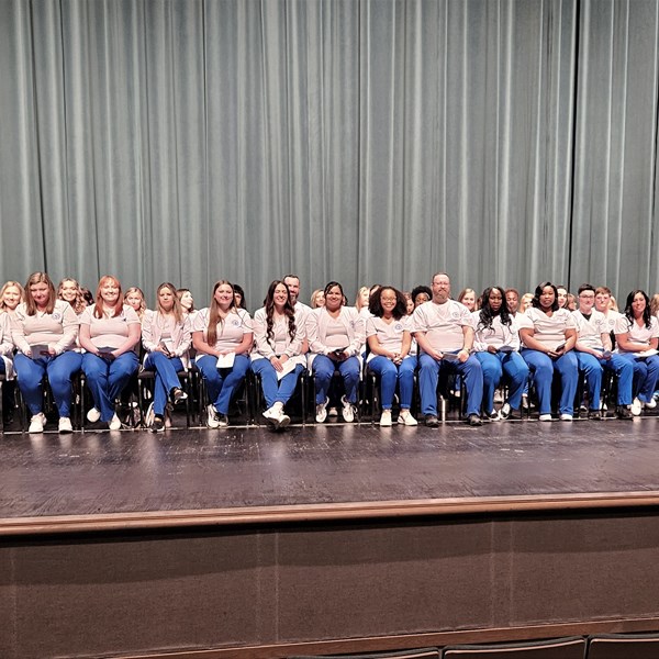 Kuss Auditorium stage with 78 nursing students sitting in chairs wearing blue scrub pants and white shirts 