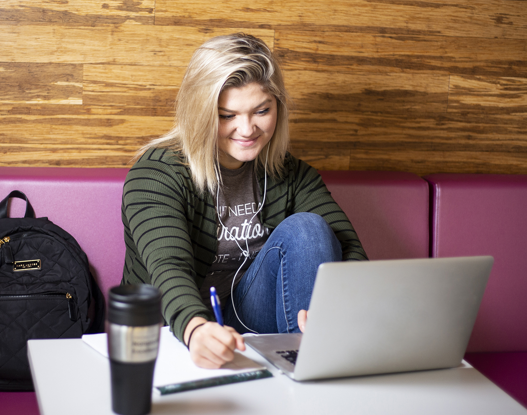 Student sitting with headphones on, looking at her laptop while taking notes.