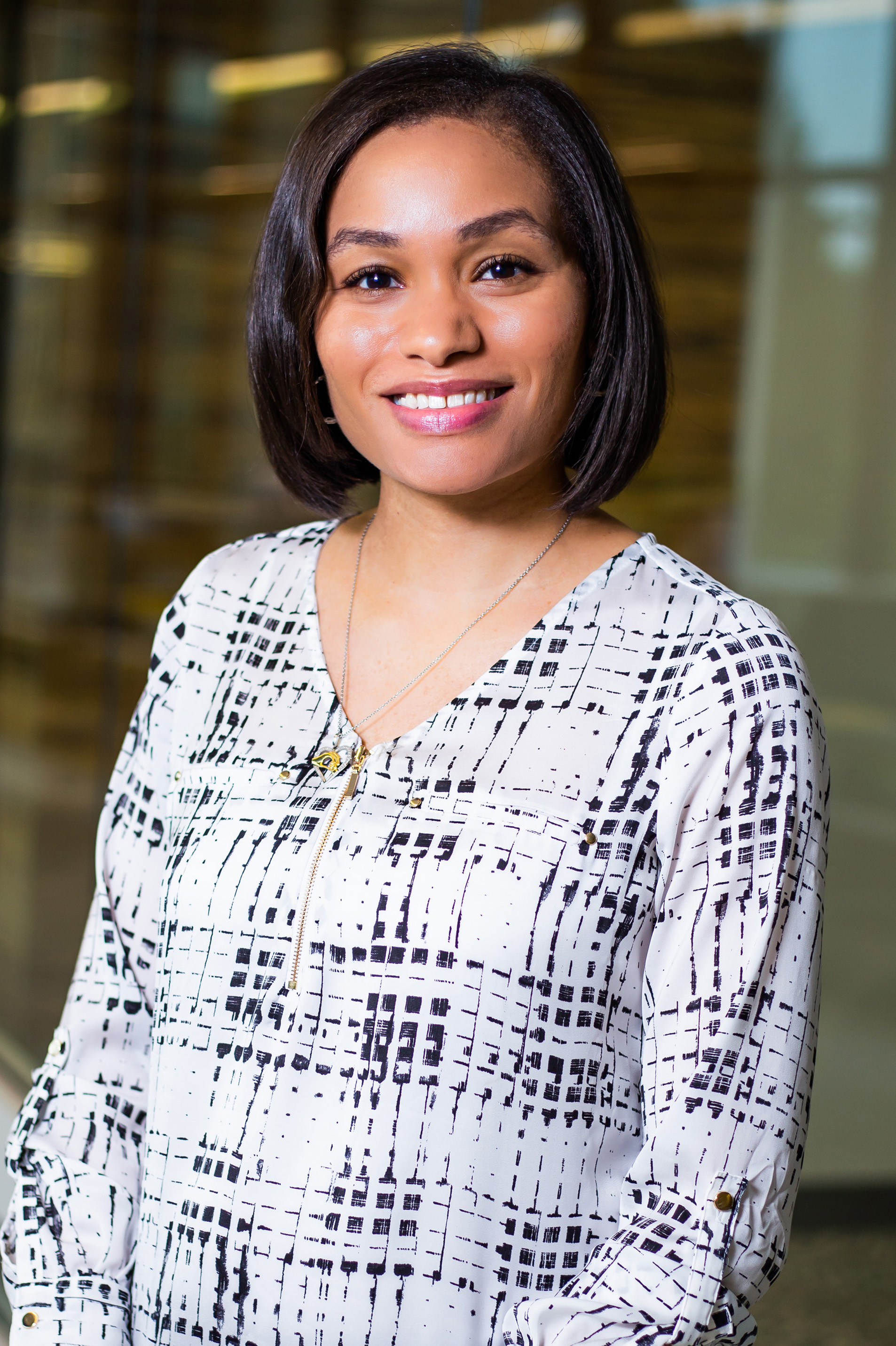Danielle Williams, a person with short brown hair wearing a black and white patterned blouse, smiling while standing indoors with a blurred glass hallway in the background.