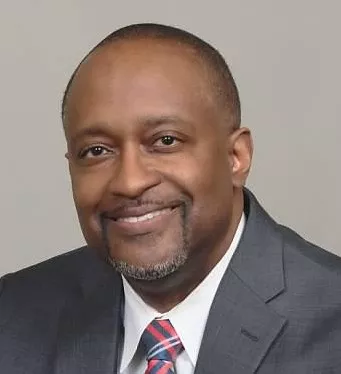African American male smiling wearing grey suit and blue and white striped tie.