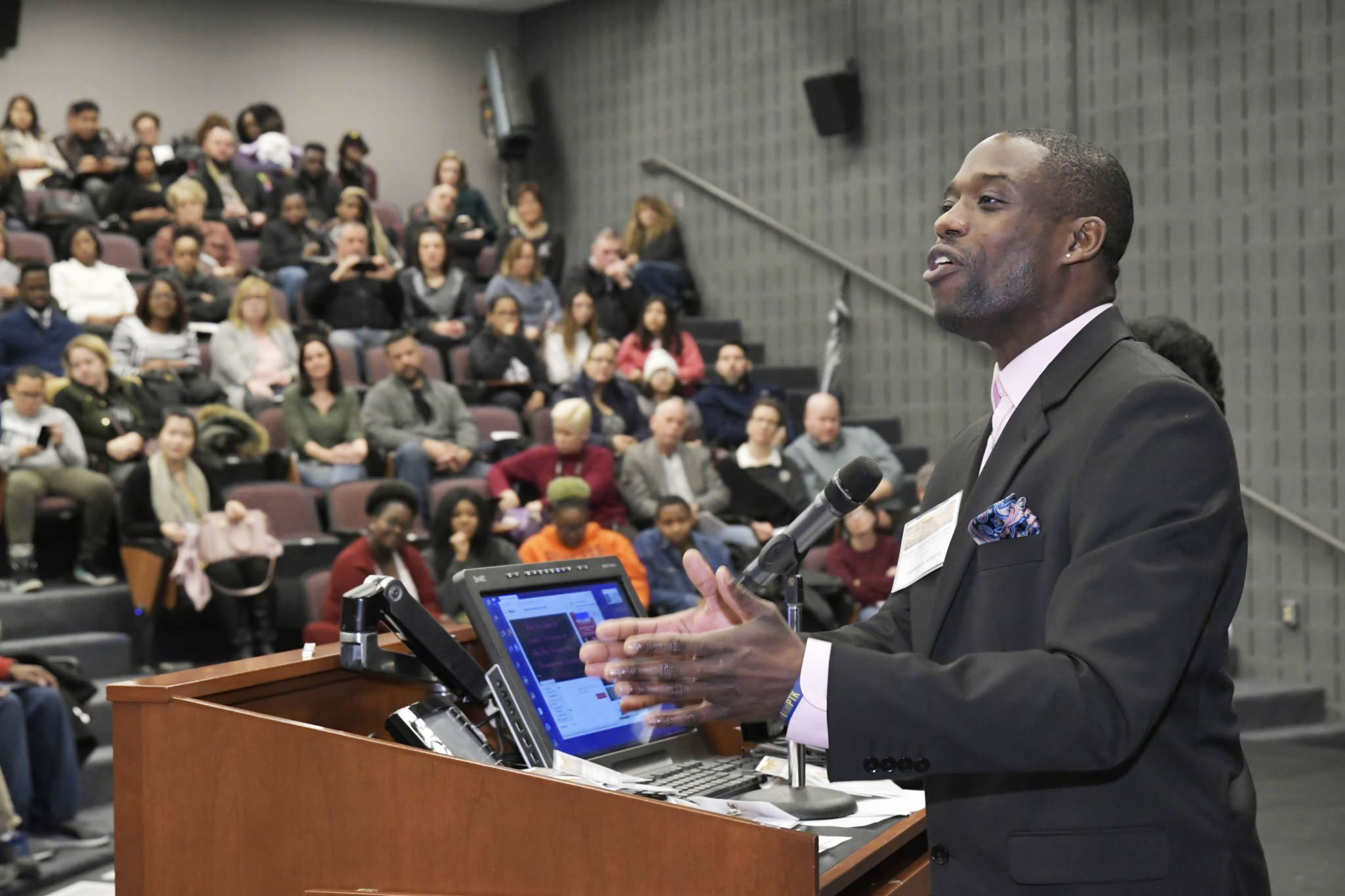African American man in dark suit stands at a podium and mic giving a presentation to a crowded room.