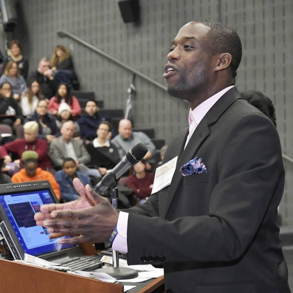 African American man in dark suit stands at a podium and mic giving a presentation to a crowded room.