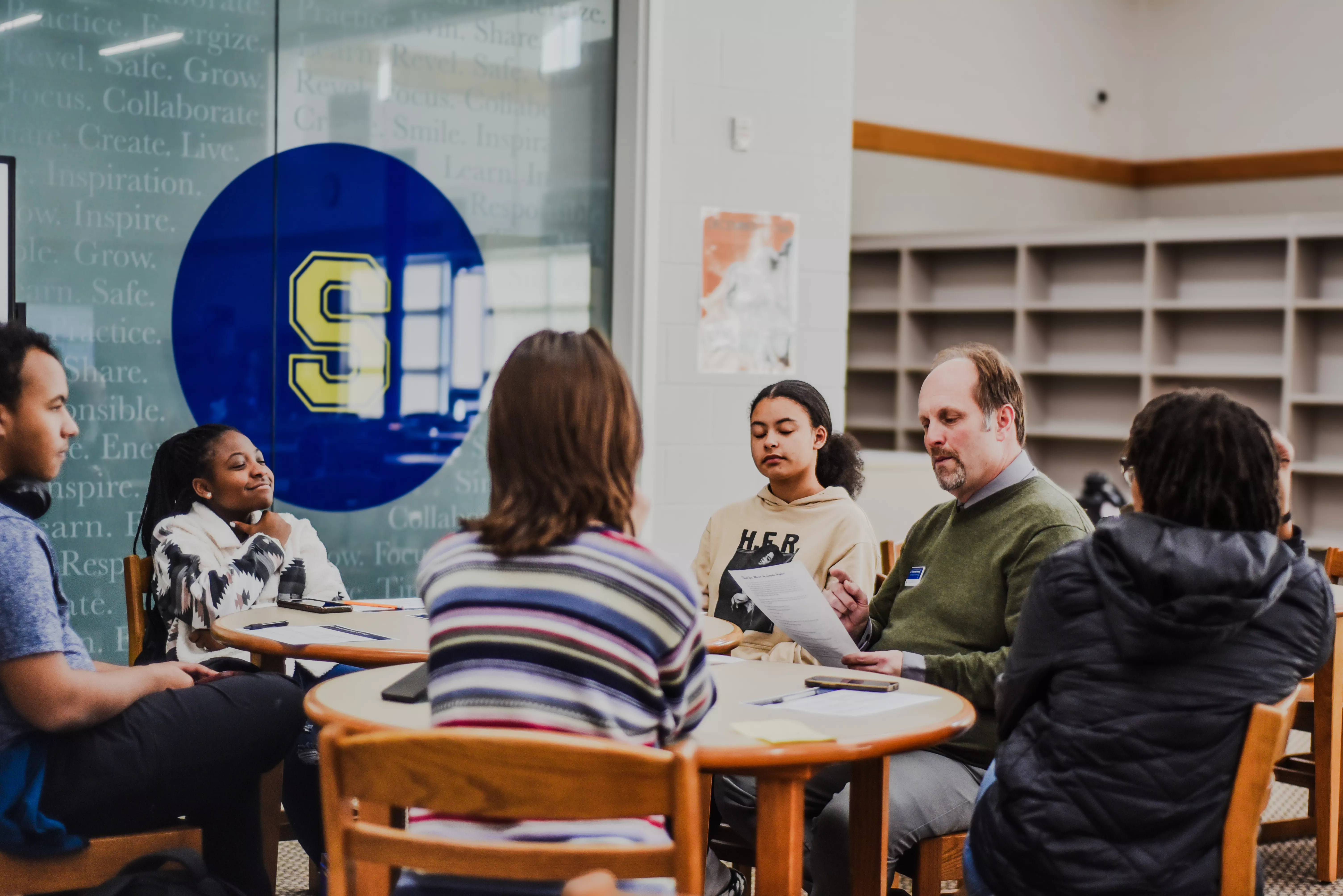 Male teacher sits at table with five students reviewing paperwork.