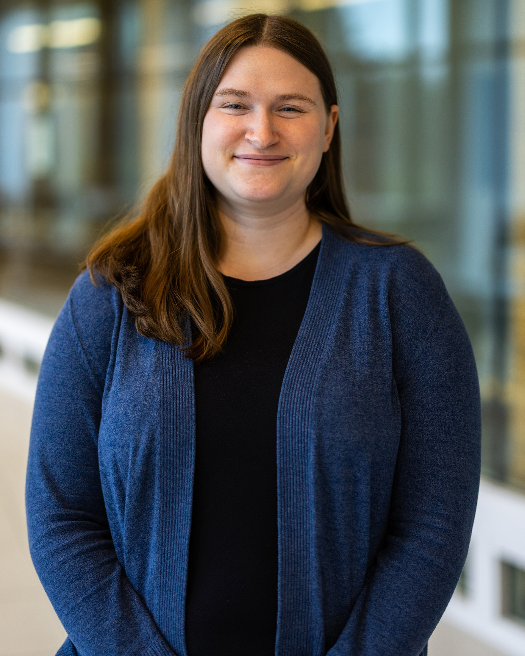 Shannon Kollasch, a person with long brown hair wearing a dark blue cardigan and black top, smiling while standing indoors with a blurred glass hallway in the background.