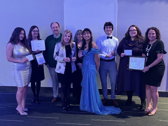 Group of nine men and women of various ages holding awards in a ballroom.