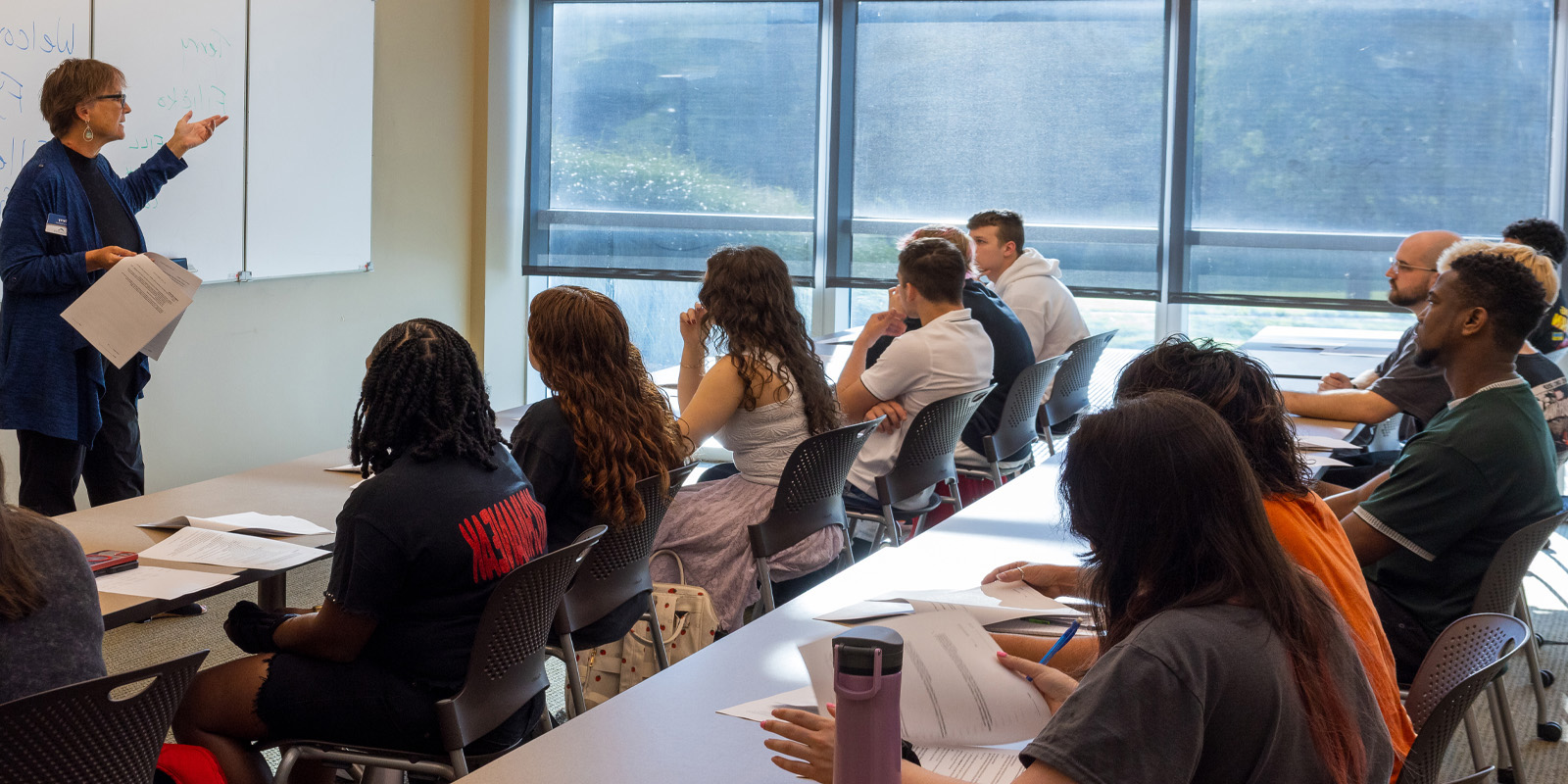 Students sitting at tables with notebooks listening to teacher at the front of the room speak.