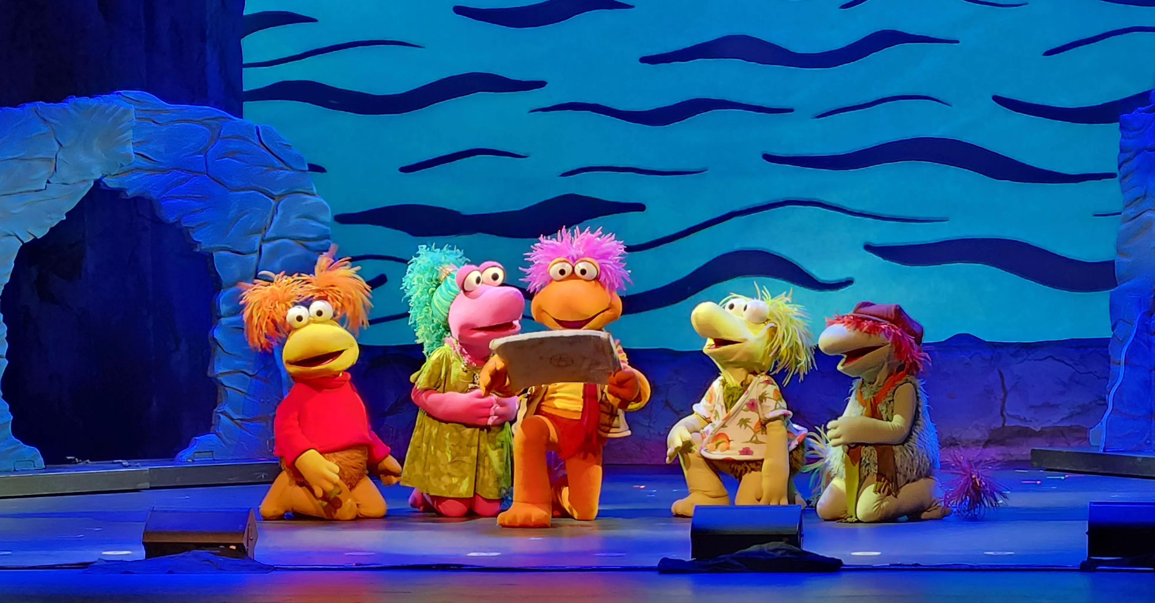 Five Fraggle Rock puppets orange, green, blue with fuzzy hair on stage with a blue background,
