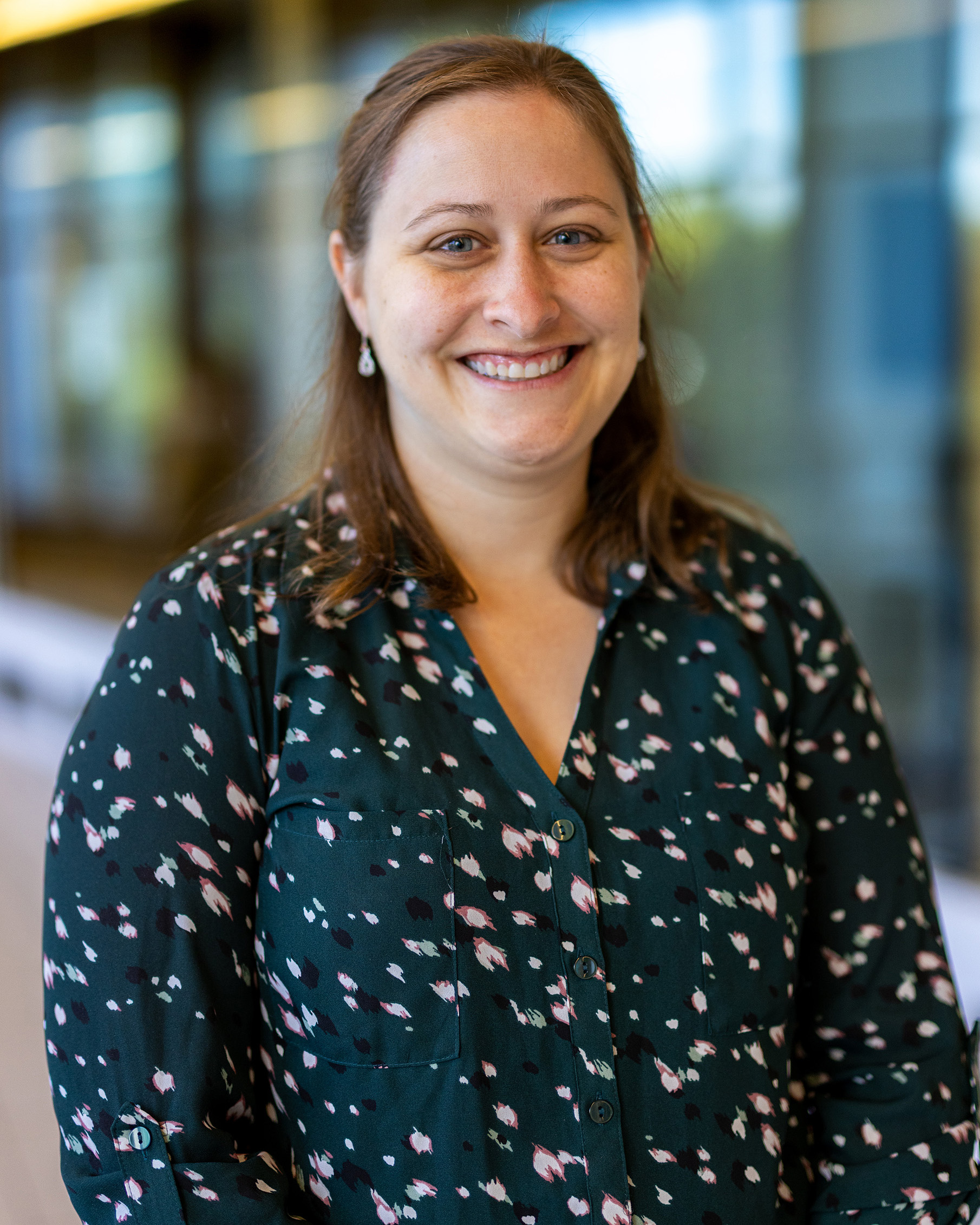 Laura McCabe, a person with light brown hair wearing a dark green patterned blouse, smiling while standing indoors with a blurred hallway and windows in the background.