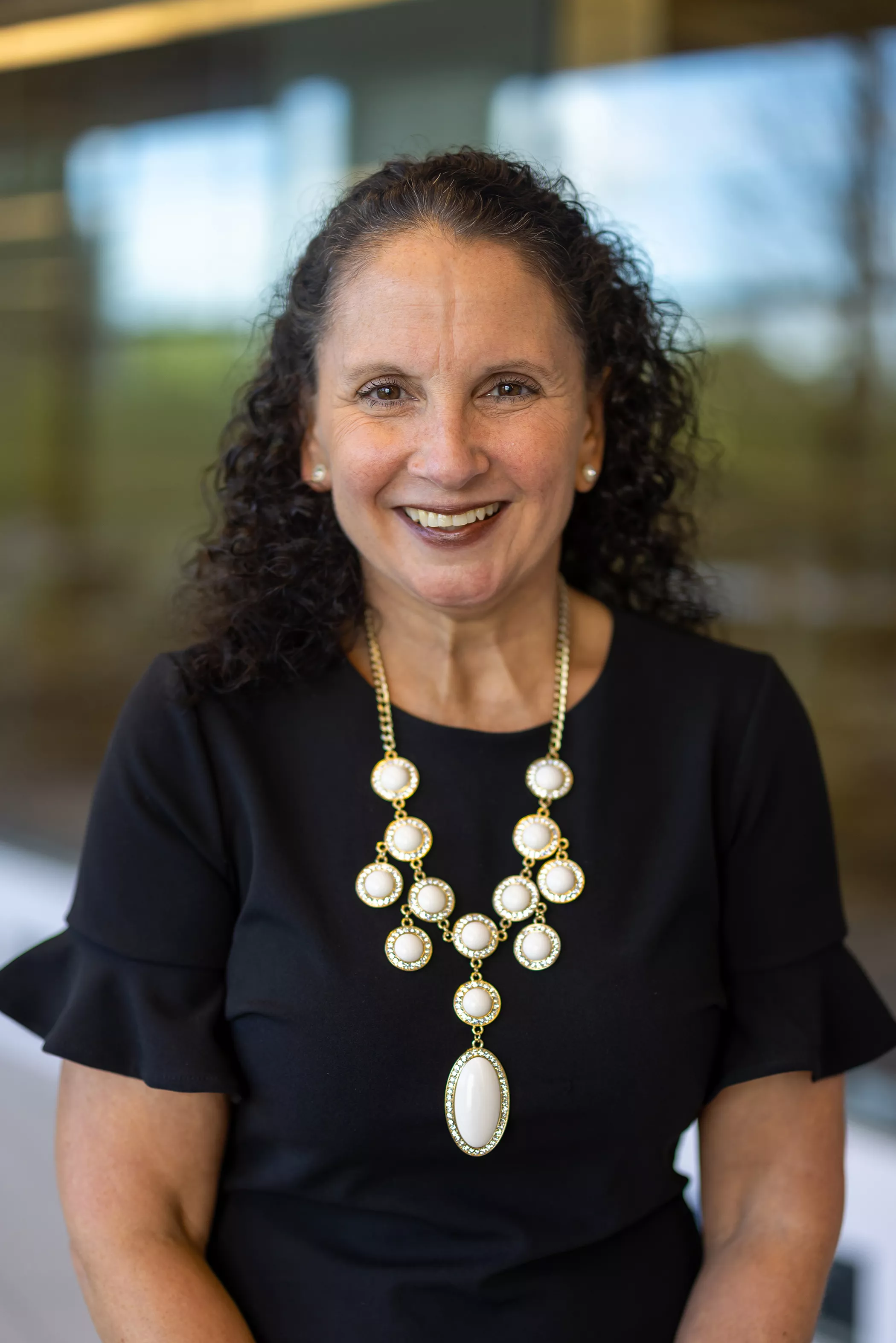 woman with dark curly hair wearing black short sleeve dress with white beaded necklace