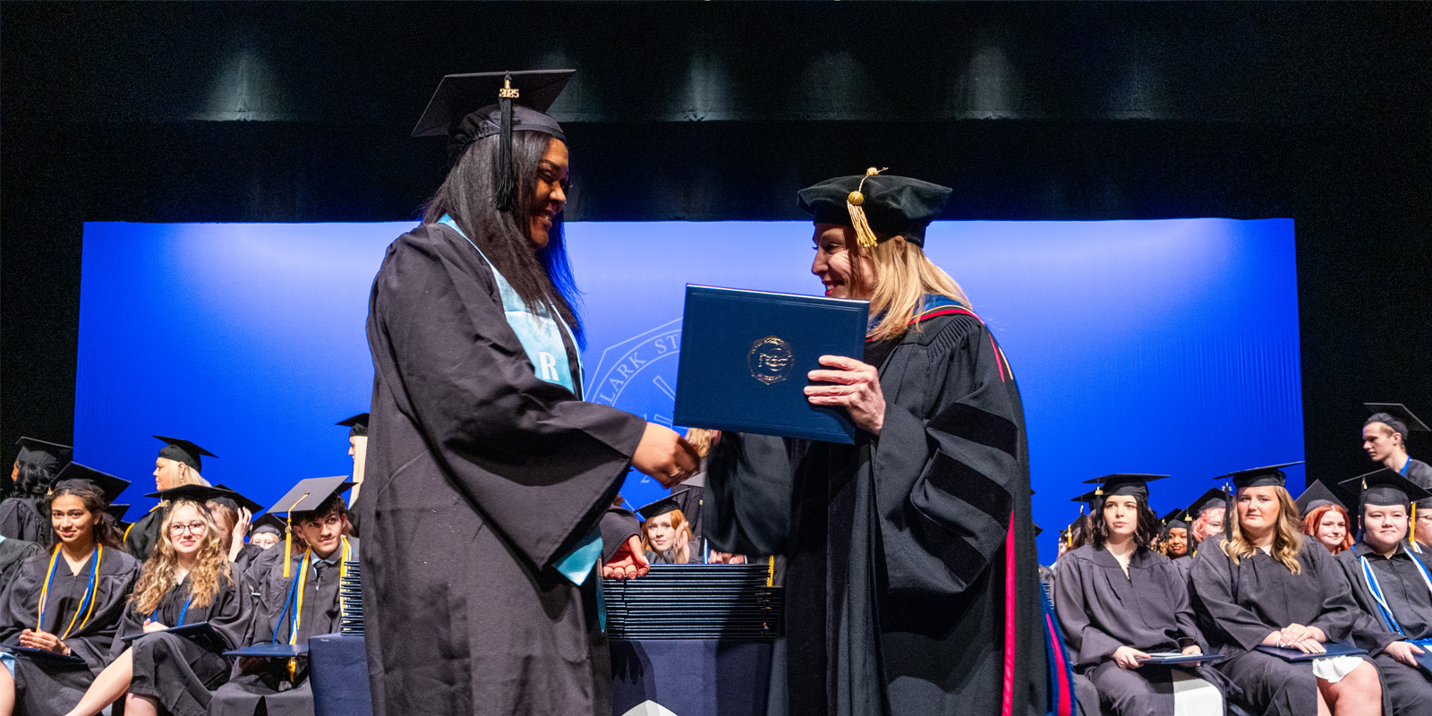 President Blondin dressed in regalia smiling and handing a diploma to a student graduating.