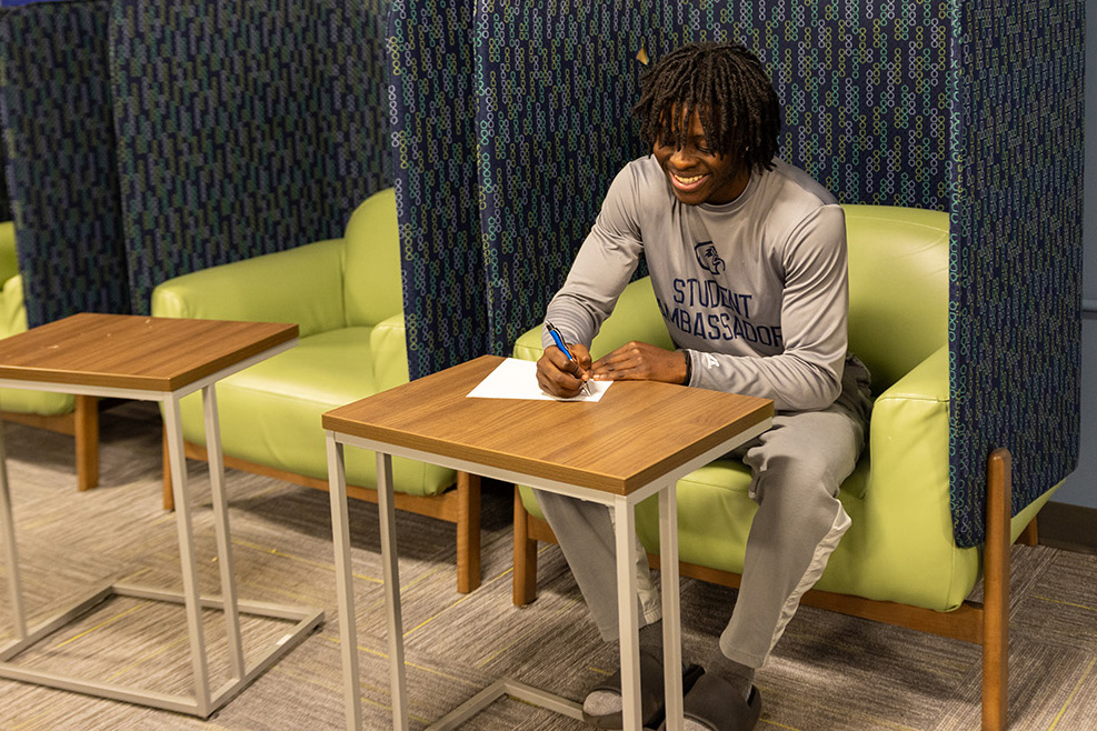 African American male student wearing a Clark State long sleeve shirt, sitting and writing in a notebook.
