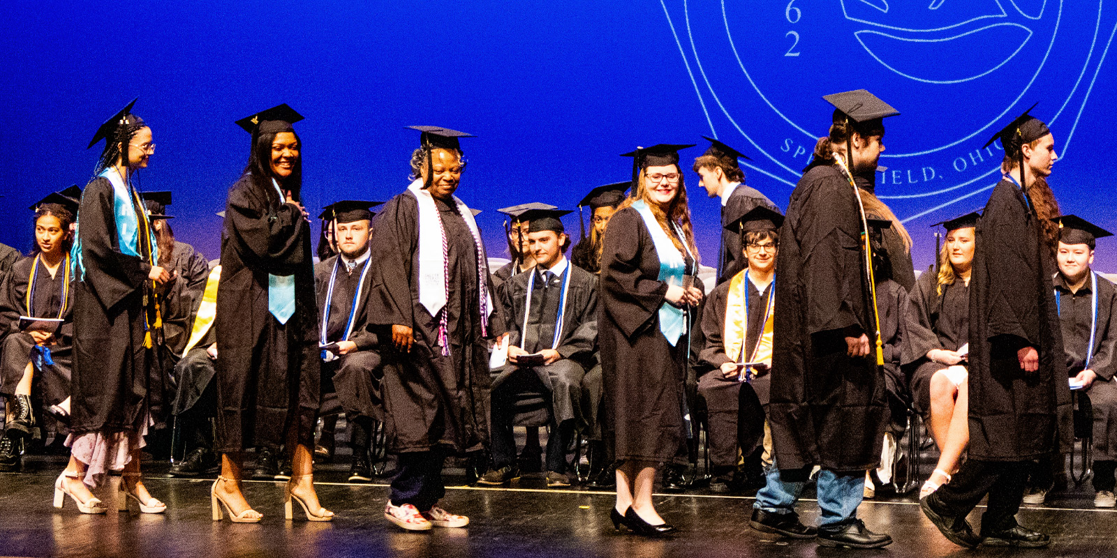 Students wearing black graduation gown and cap walking across the stage at graduation.