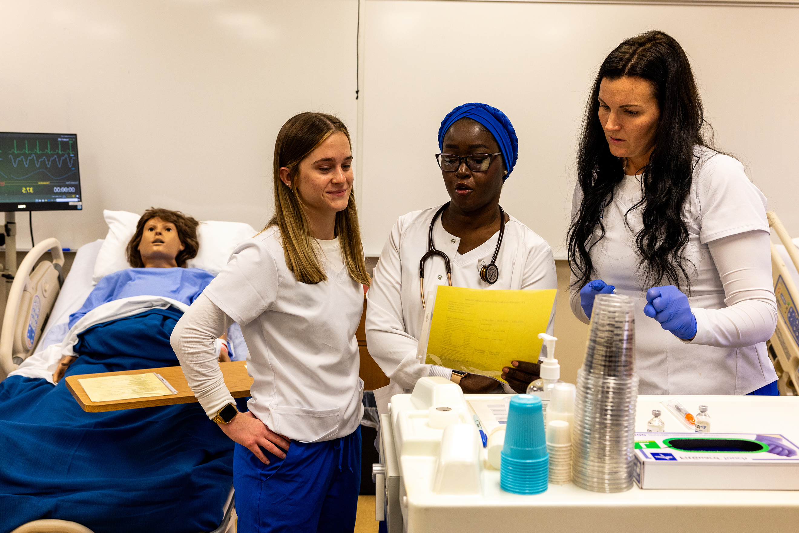 Two healthcare students in scrubs scan a wristband barcode beside a laptop.