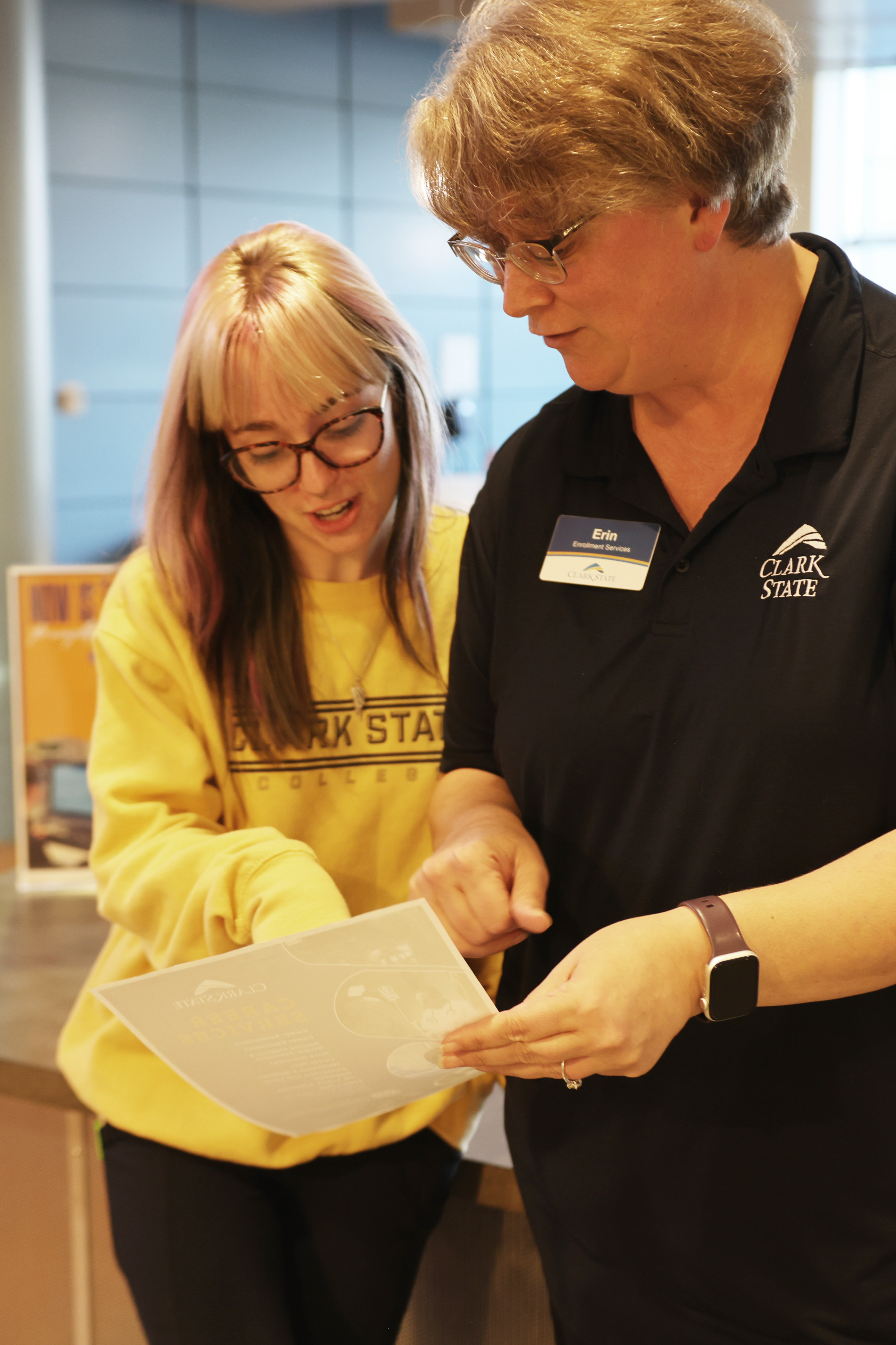 A student wearing a yellow Clark State sweatshirt pointing at a paper a staff member holds.