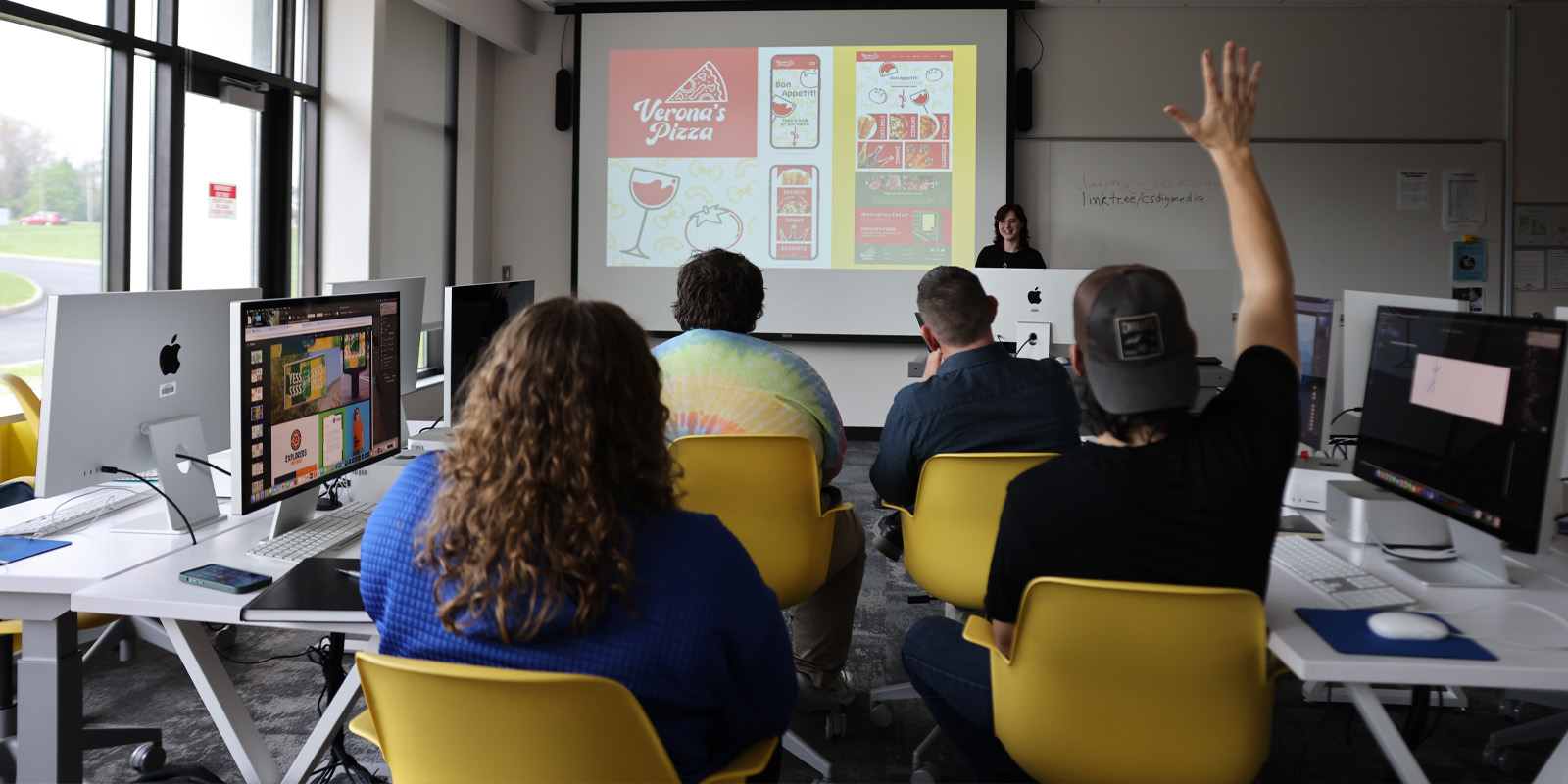 Students sitting in yellow chairs listening to a student present their design project.