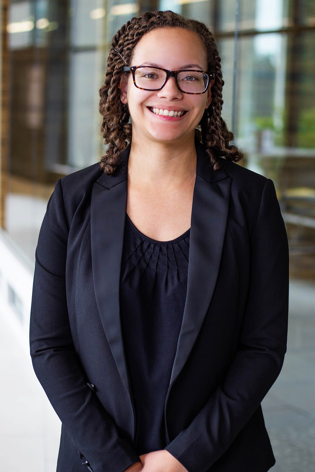 Nicole Trimble, a person with curly hair wearing glasses, a black blouse, and a black blazer, smiling while standing indoors with a blurred glass hallway in the background.