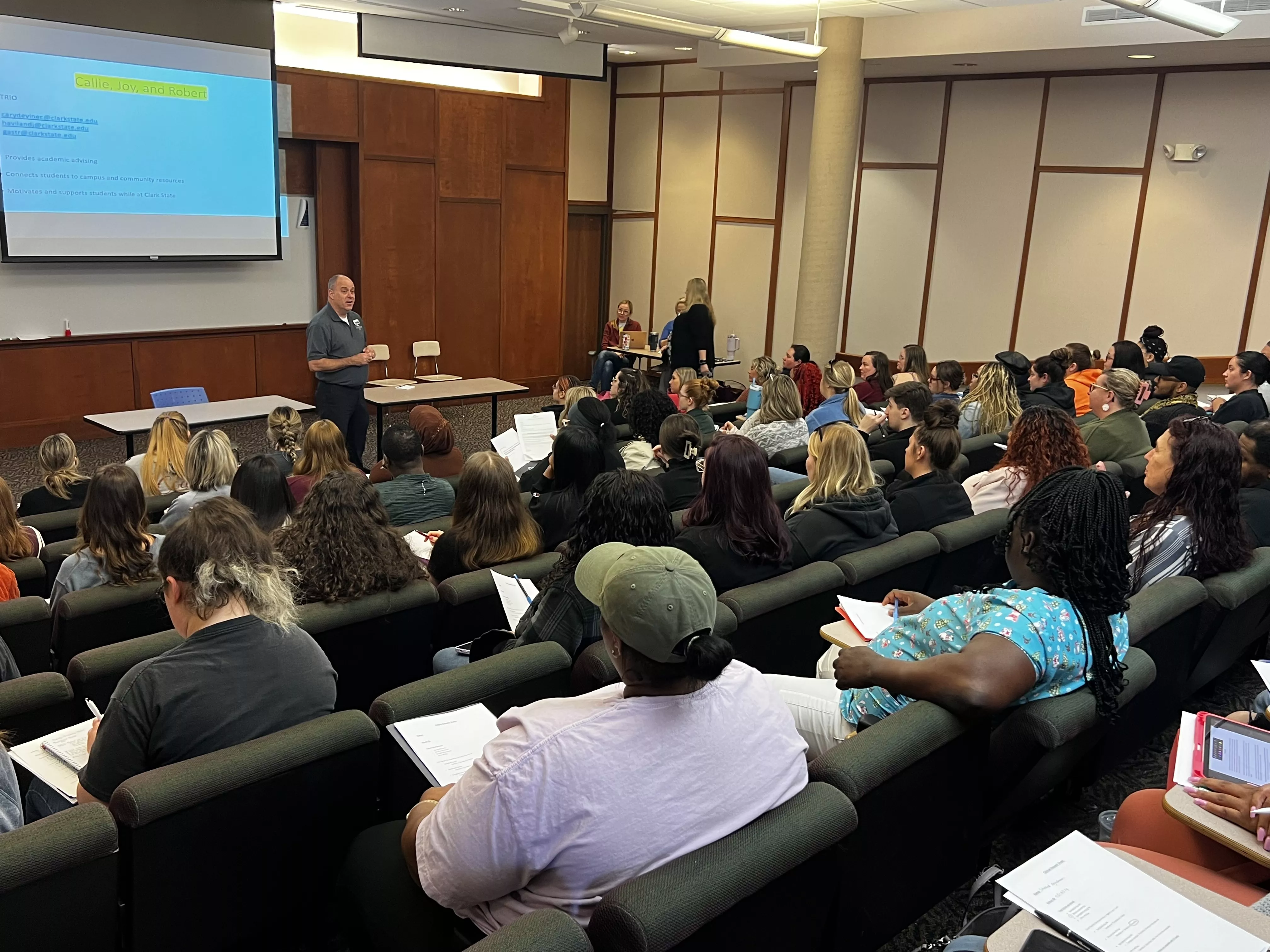 Classroom with green chairs and 50 students watching a screen and man giving a presentation.