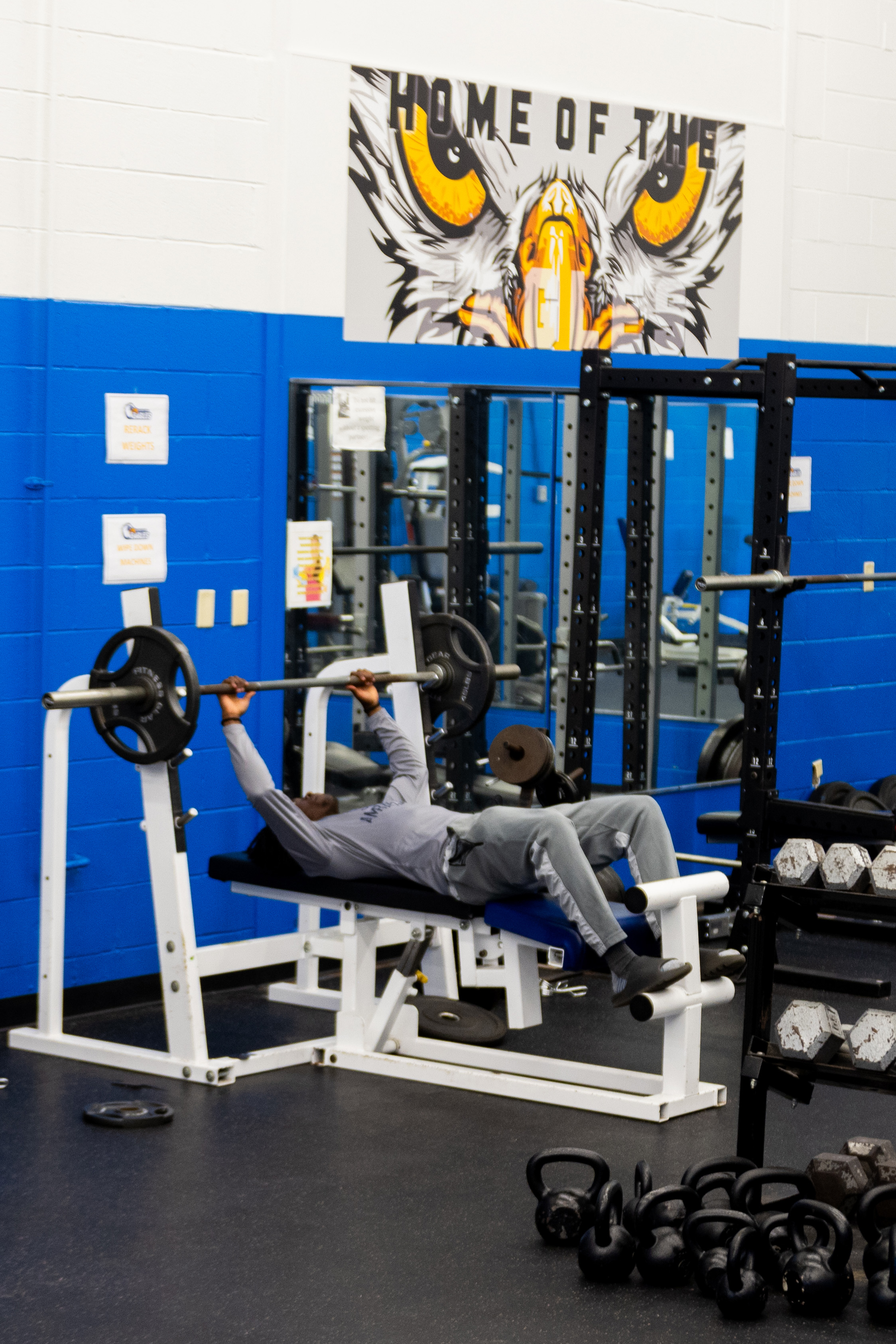 A person lies on a bench press in a campus gym, lifting a barbell, with weight racks, kettlebells, and an eagle mural reading “Home of the Eagles” in the background.