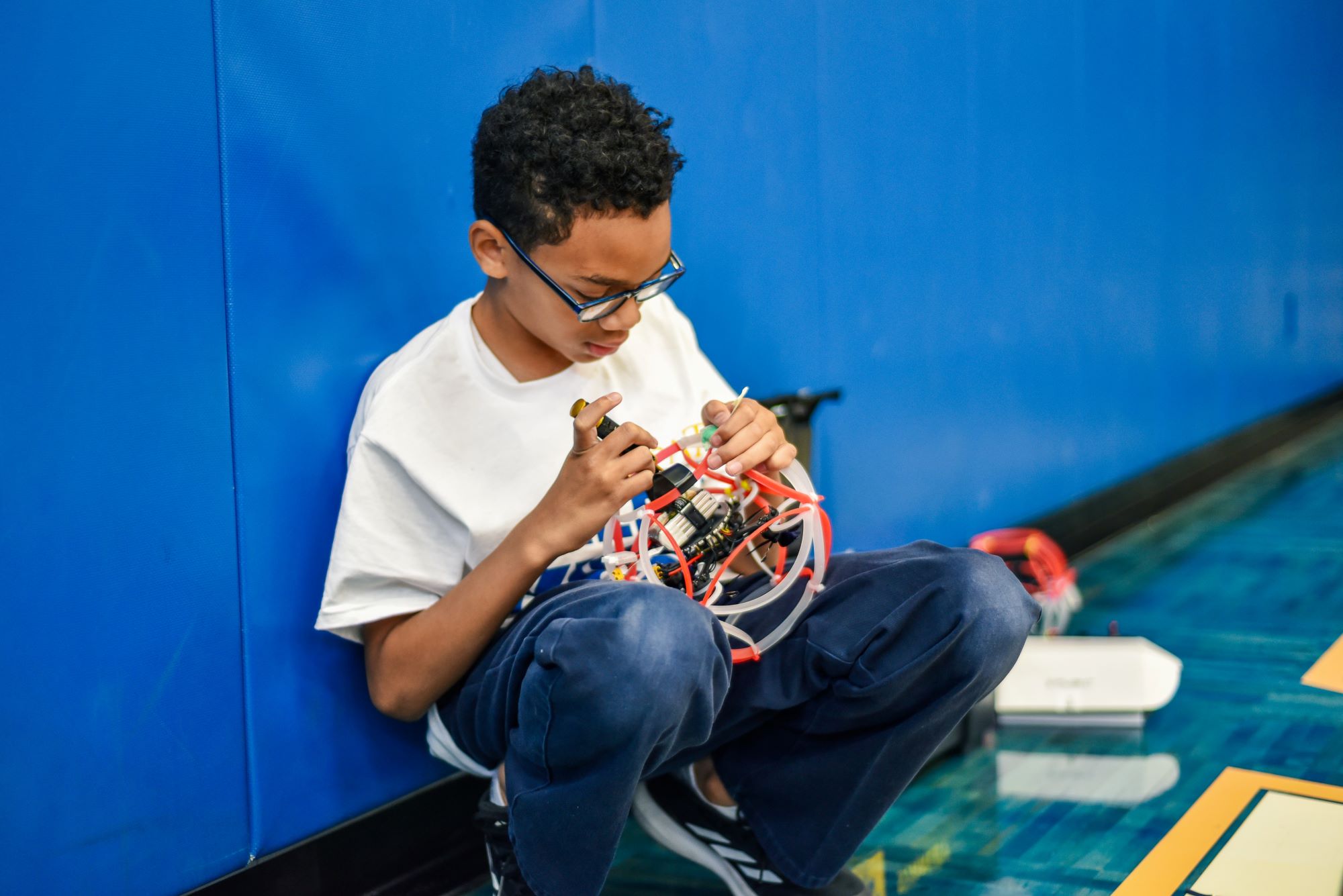 Summer camp student in white t-shirt against blue wall builds a drone soccer ball.