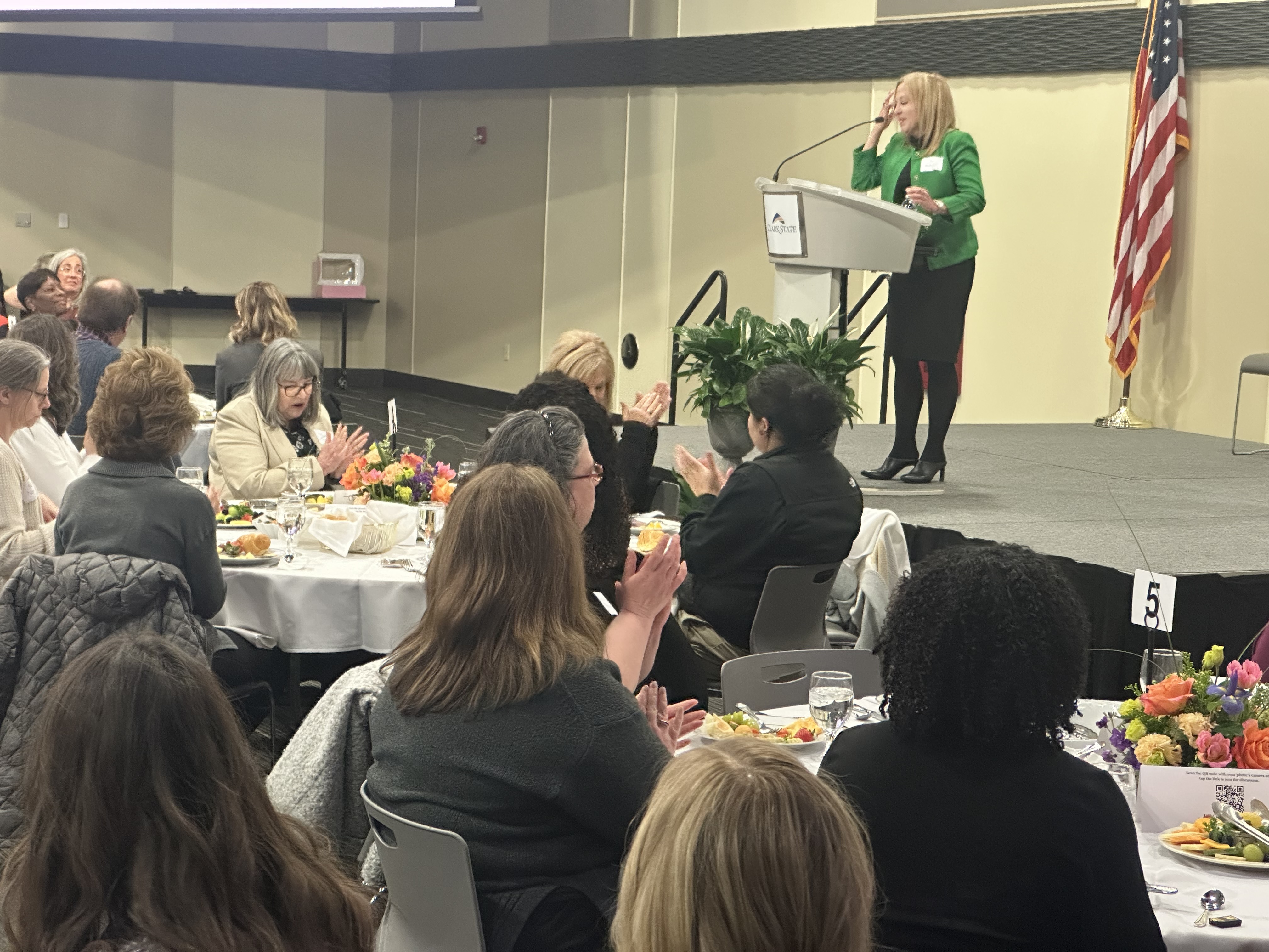 Lunch ball room with woman in green jacket standing at podium with audience eating lunch.