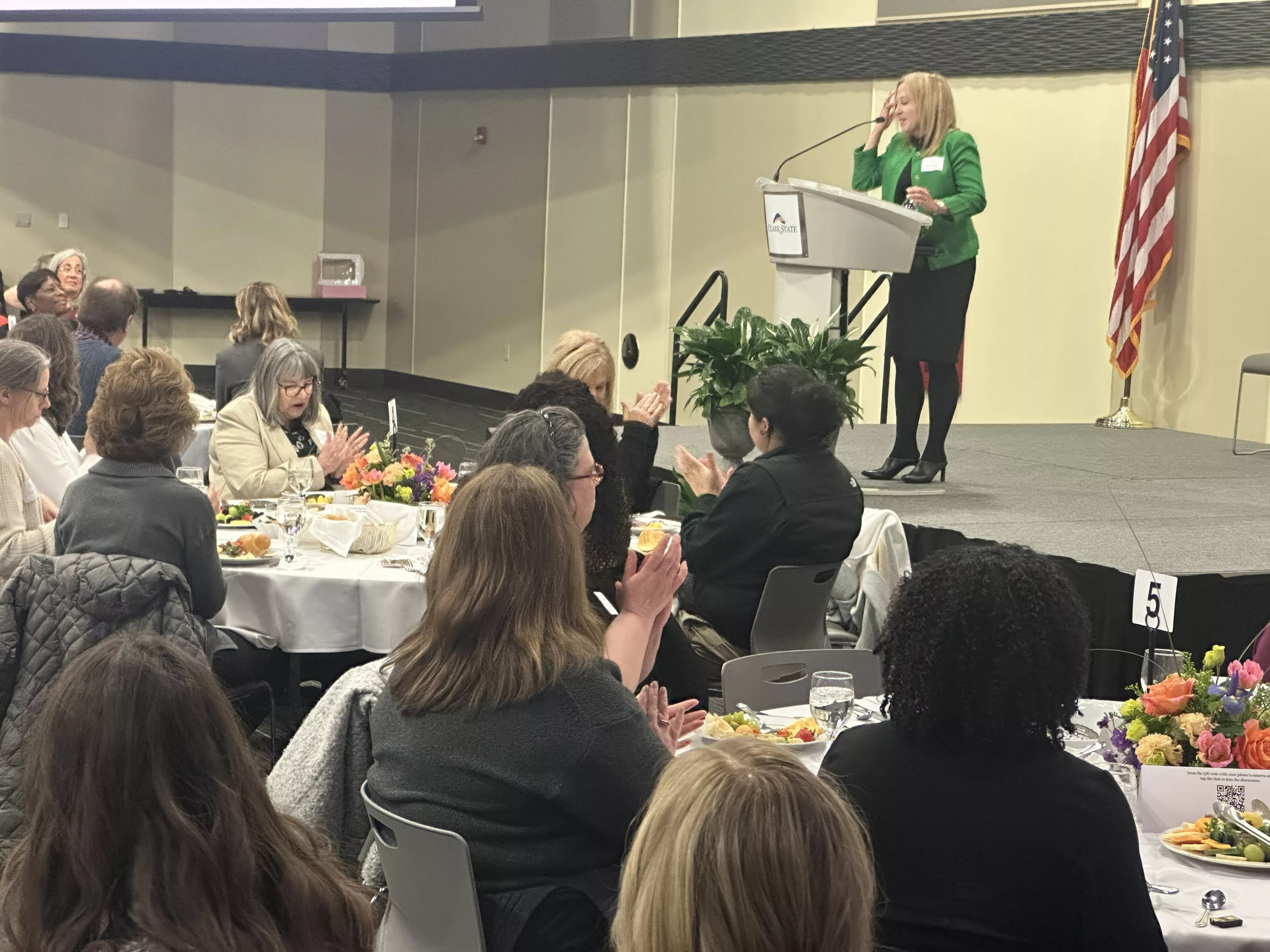 Lunch ball room with woman in green jacket standing at podium with audience eating lunch.