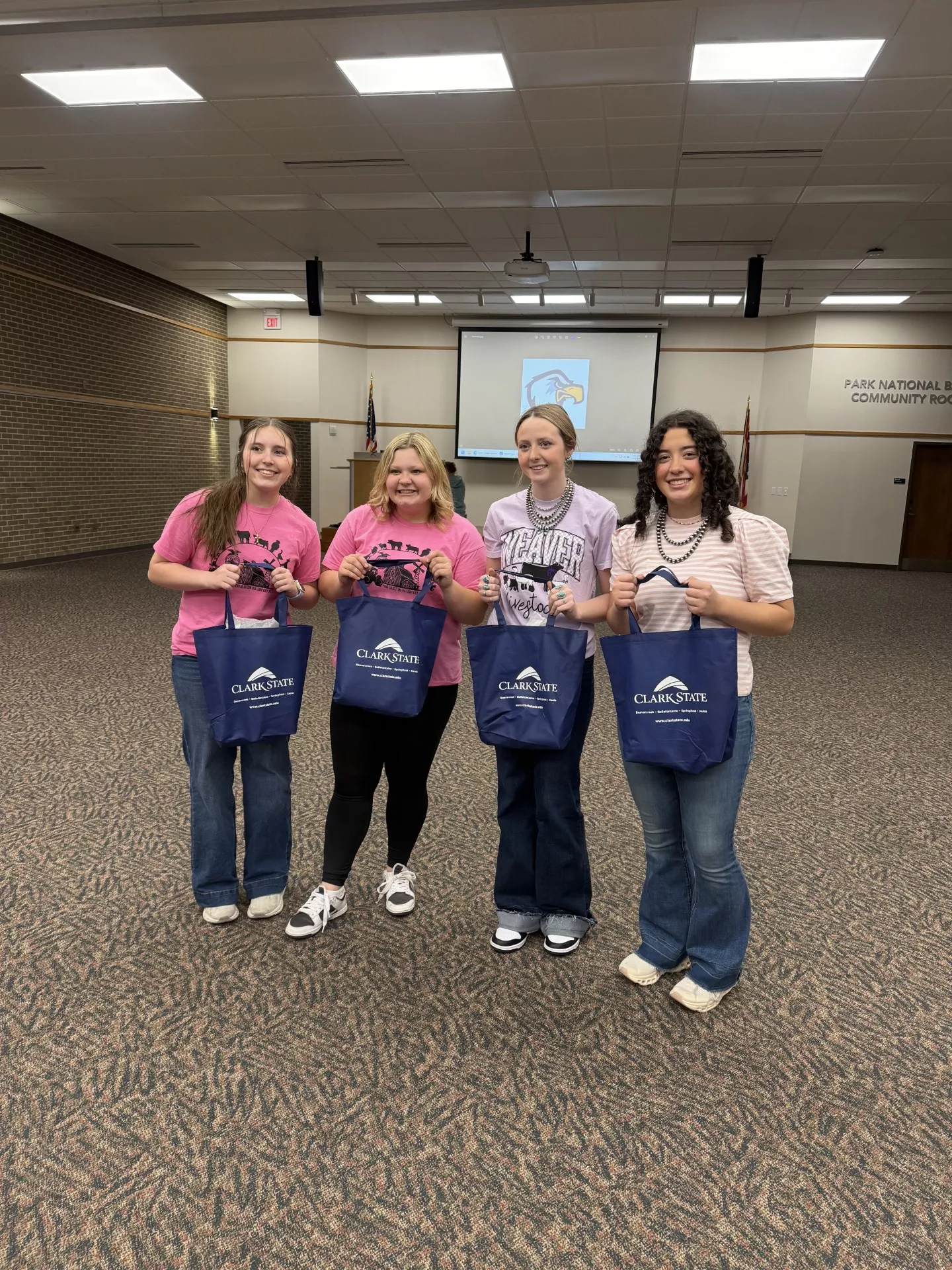 Four smiling girls wearing pink shirts, jeans and holding blue Clark State bags.