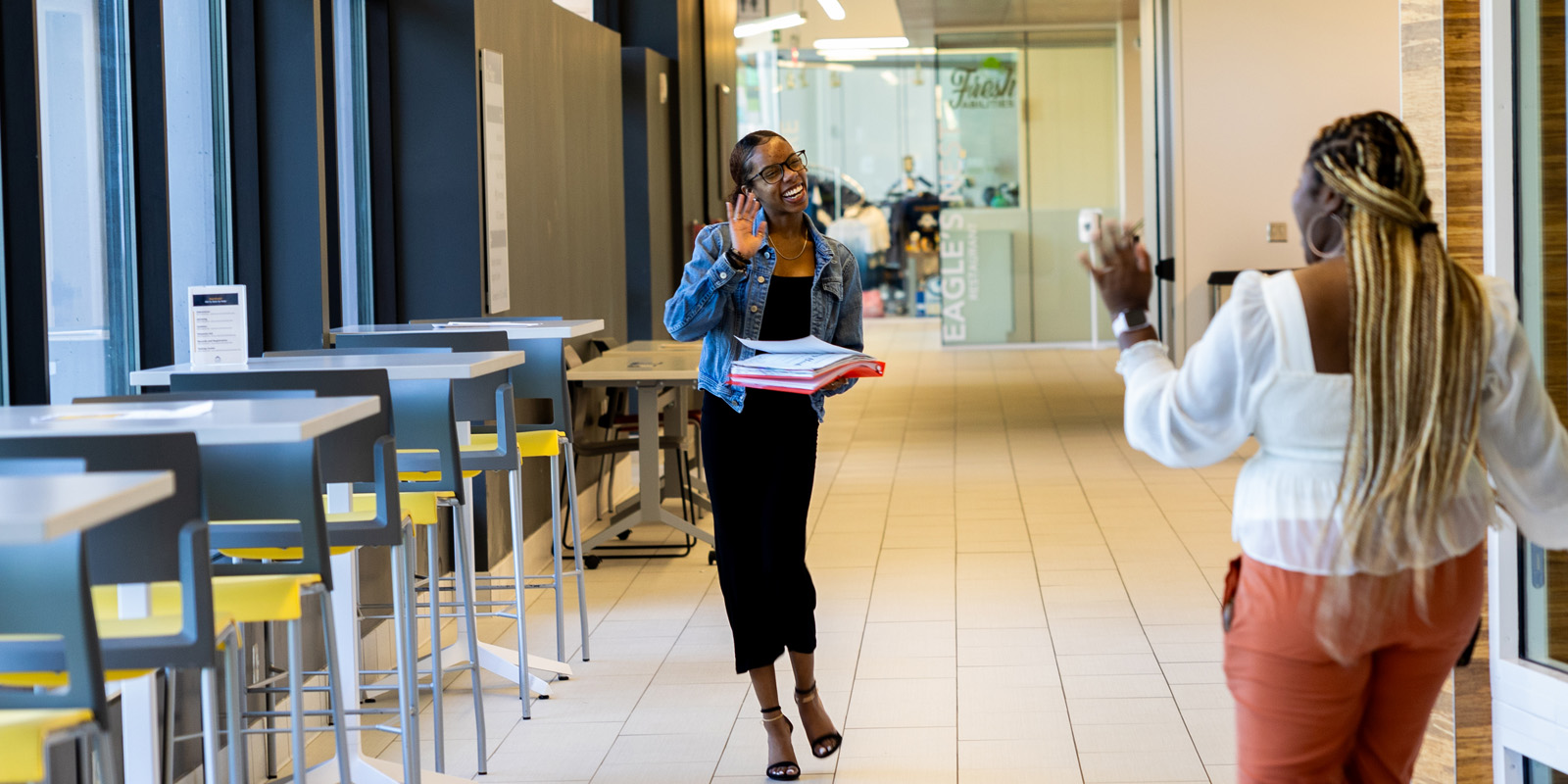 African american female student wearing a black dress and jean jacket waving to another student in a hallway.
