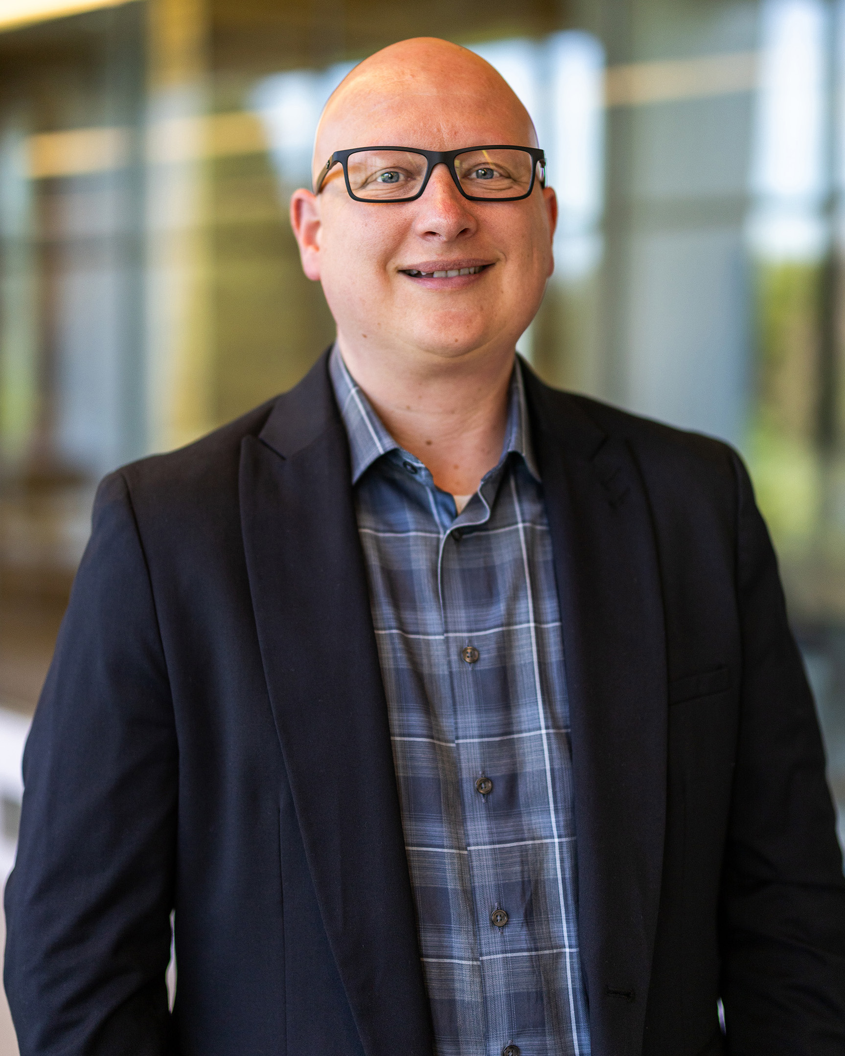 Nicolas Sparrow, a person with no hair wearing glasses, a blue paid shirt, and a black blazer, smiling while standing indoors with a blurred glass hallway in the background.