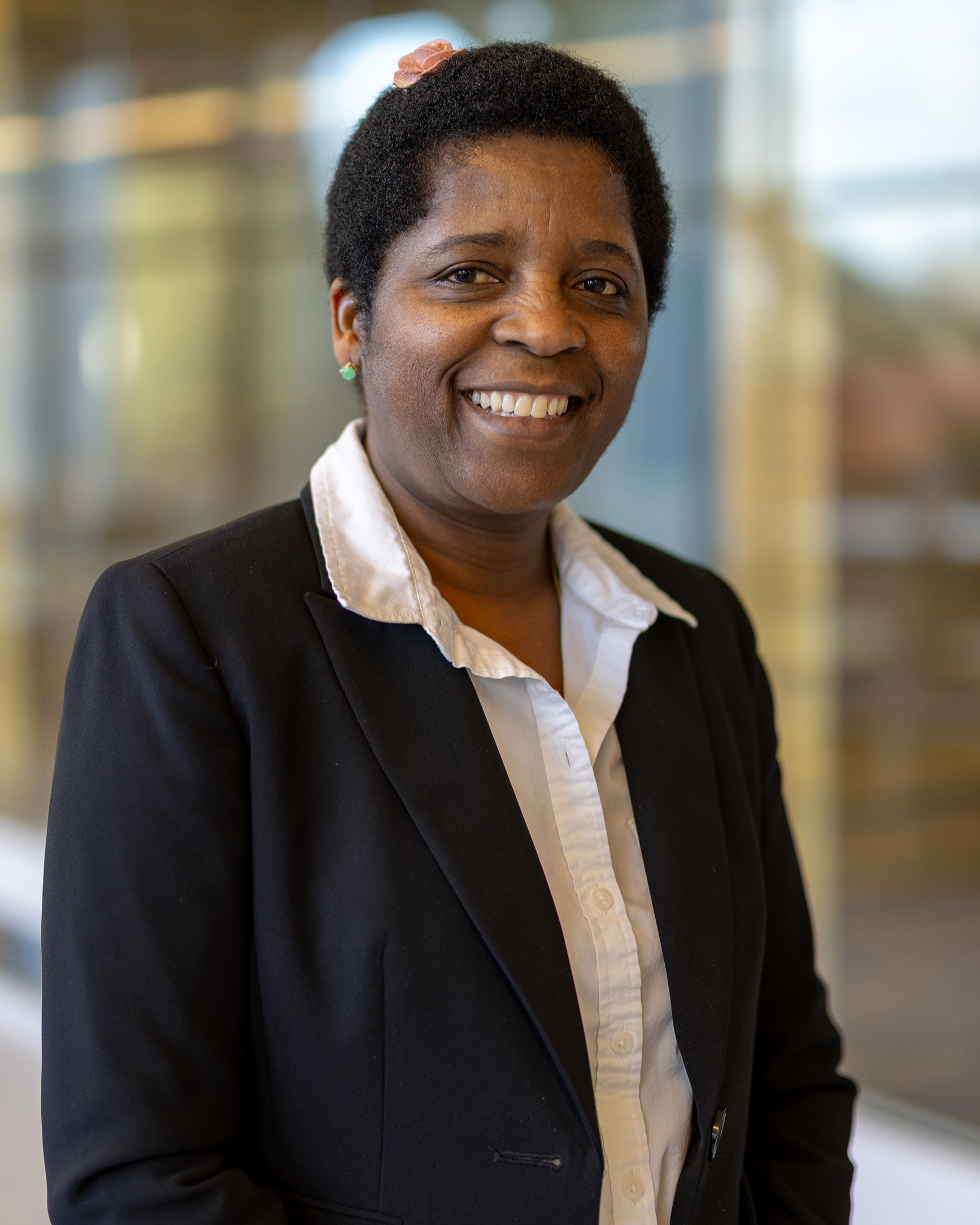 Joy Haviland, a person with curly hair, a white shirt, and a black blazer, smiling while standing indoors with a blurred glass hallway in the background.
