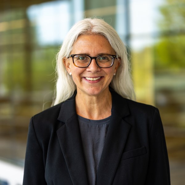 Woman with long grey hair and glasses wearing navy blue suit jacket.