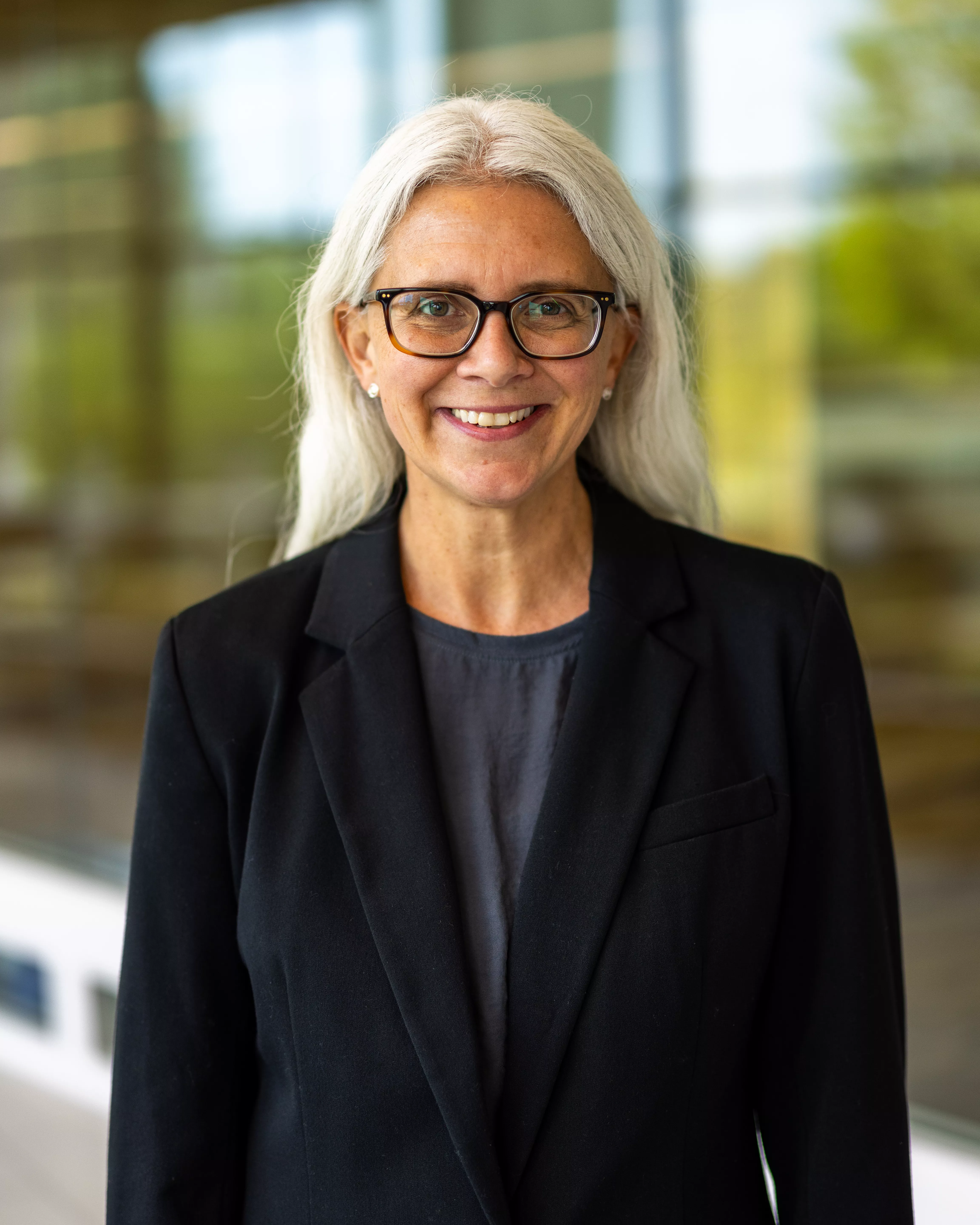 Woman with long grey hair and glasses wearing navy blue suit jacket.