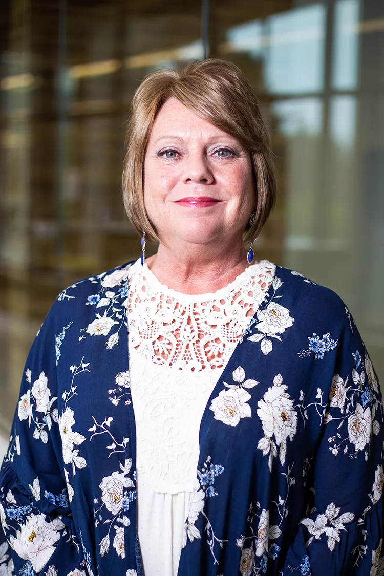 Headshot of a woman with brown hair wearing a navy blue jacket with white flowers.