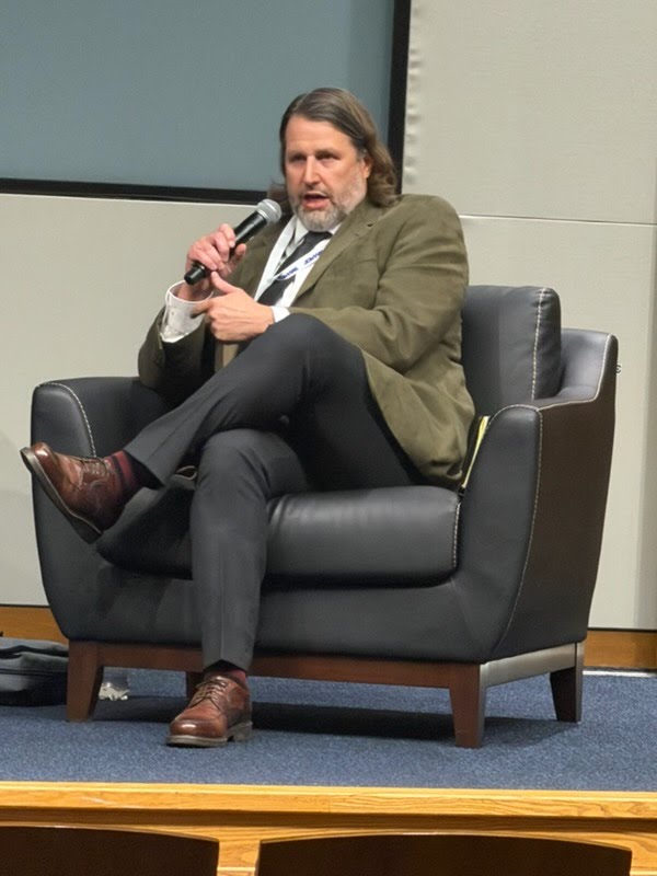 Man sitting in blue chair with microphone wearing tan sport jacket.