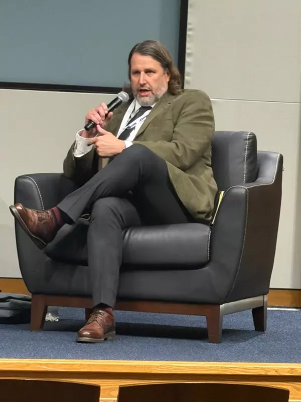 Man sitting in blue chair with microphone wearing tan sport jacket.