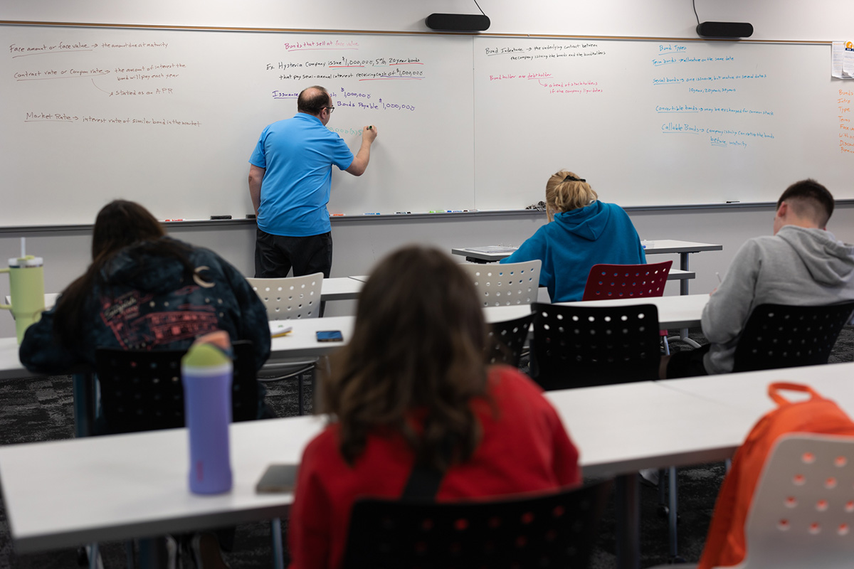 Students sitting at tables listening to faculty present at whiteboard at front of classroom.