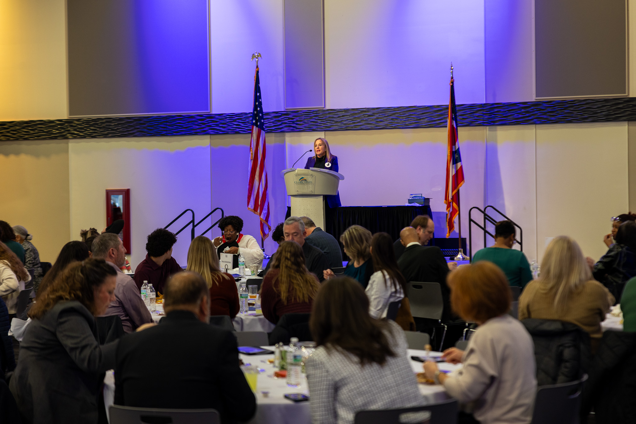 Woman standing at podium with audience seated at tables in front of the stage.
