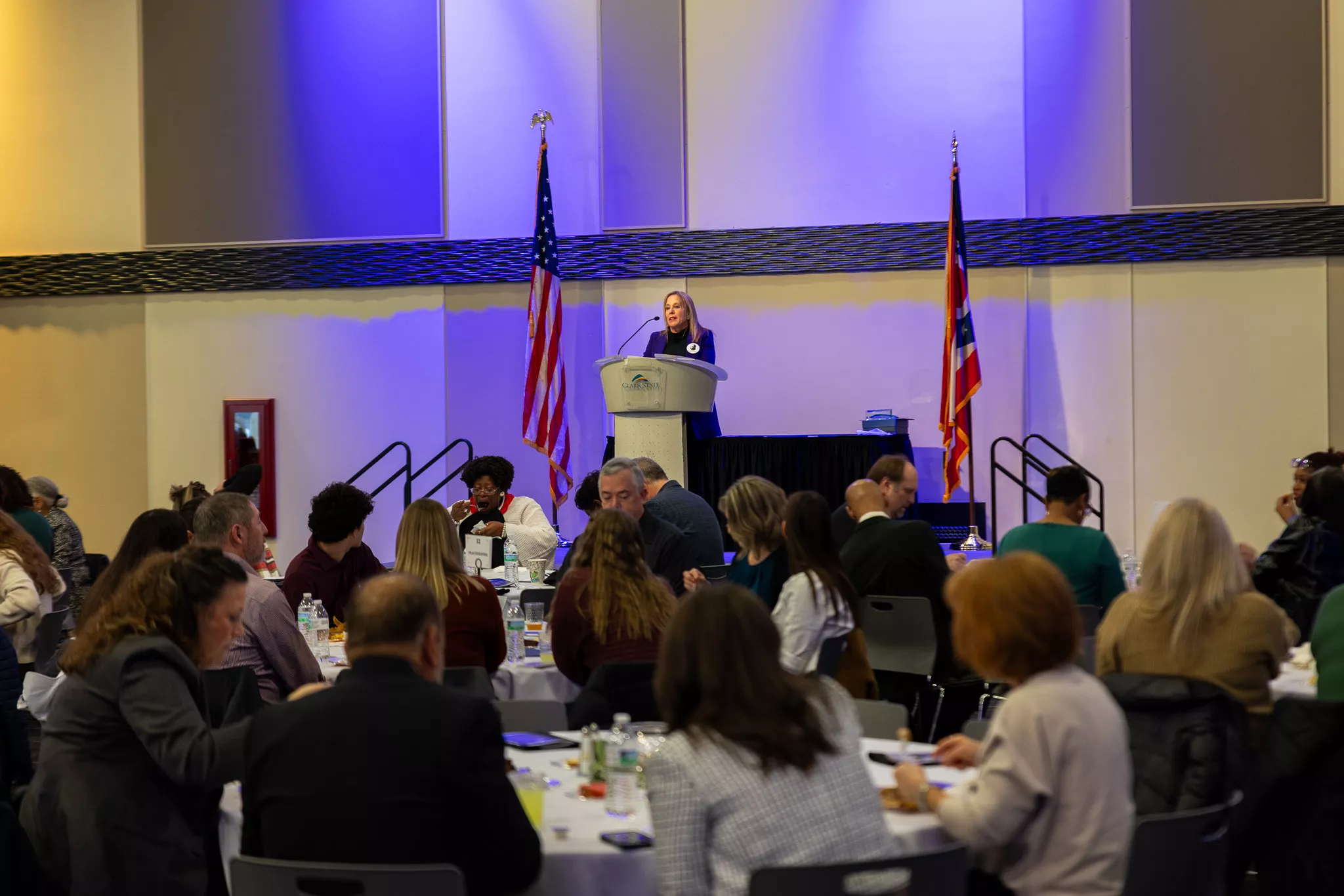 Woman standing at podium with audience seated at tables in front of the stage.