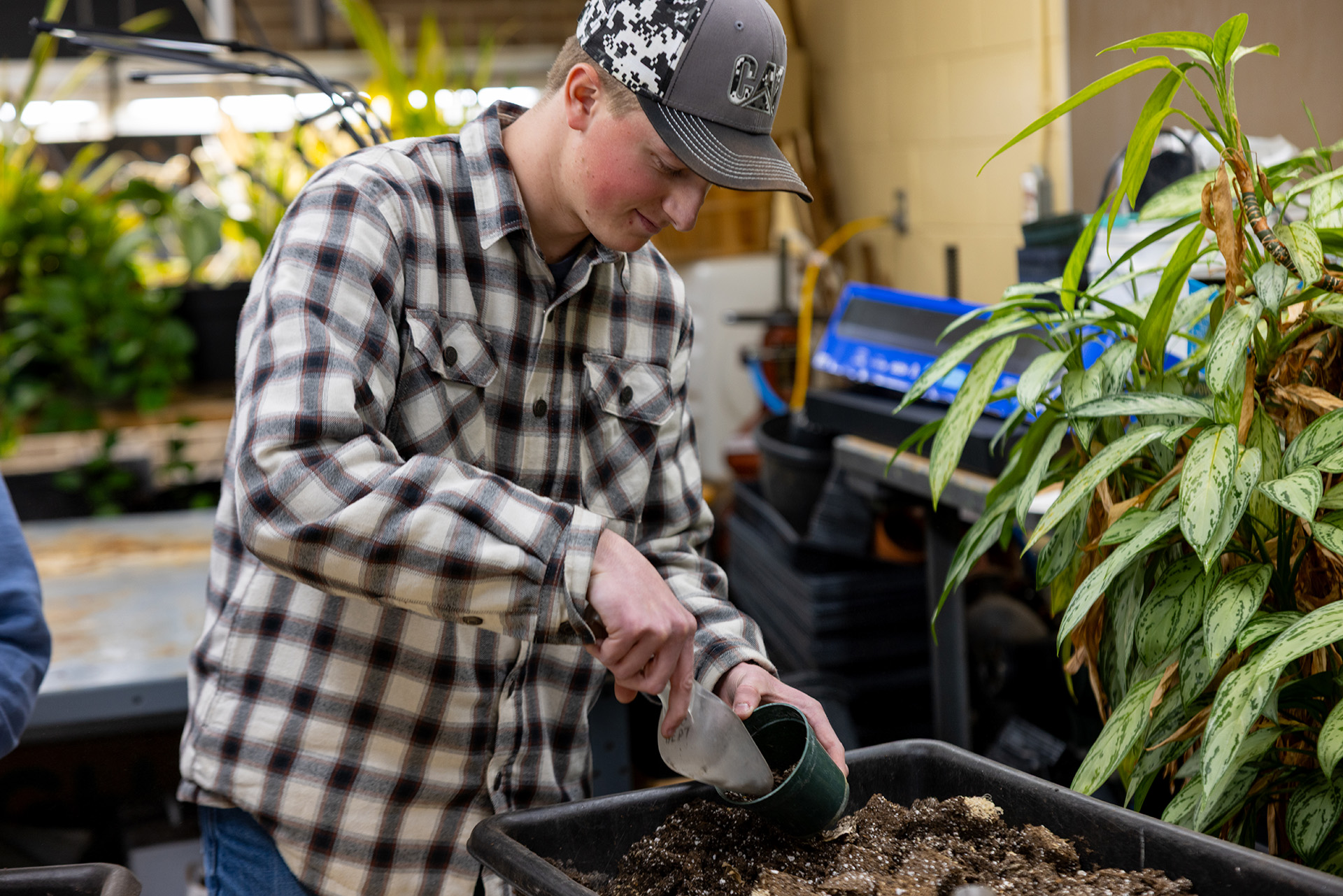 Student wearing a ball cap and plaid shirt using a trowel to put dirt into a plastic pot.