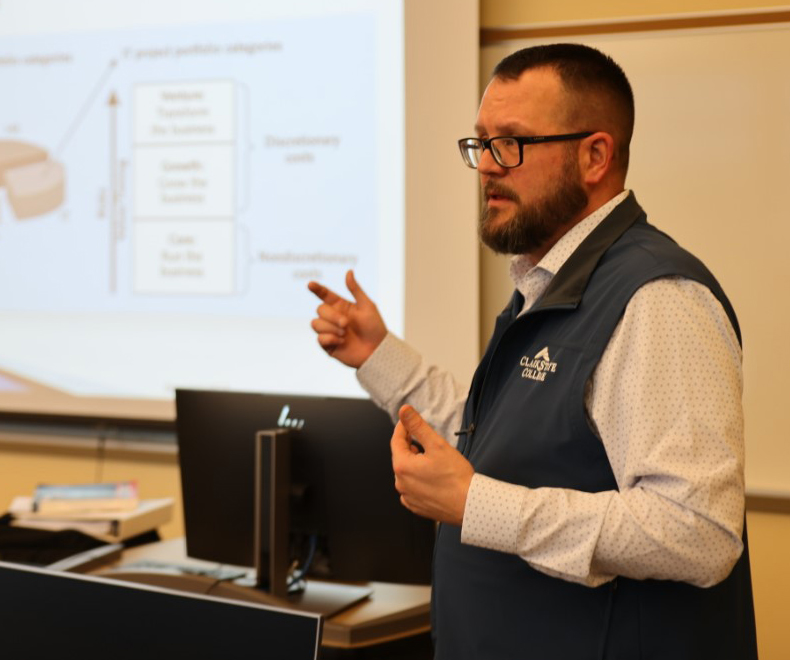 Instructor stands at front of classroom showing a chart about stocks on projector screen.
