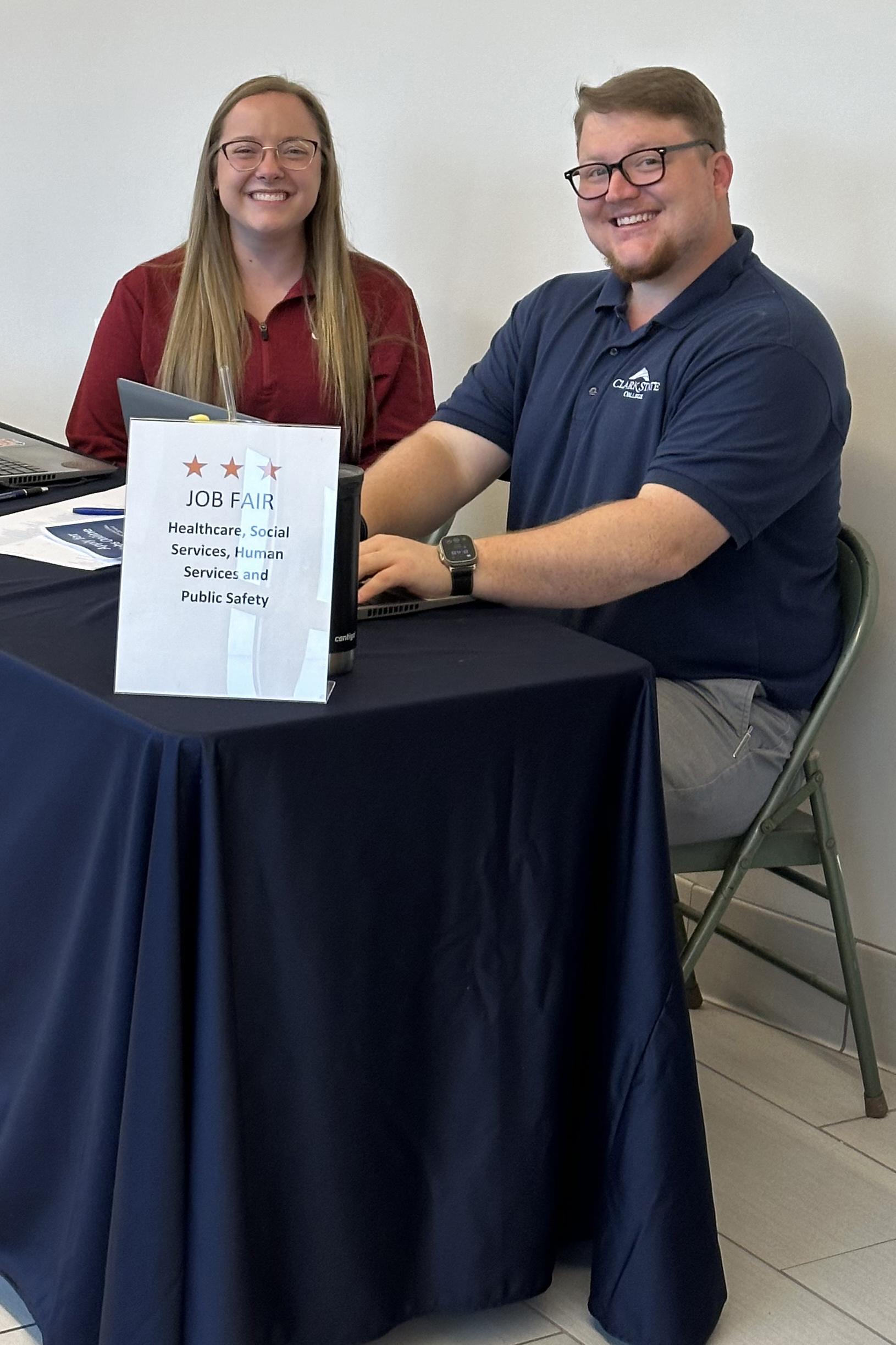 Two smiling staff members sit behind a table at a job fair with laptops and a sign reading “Job Fair.”