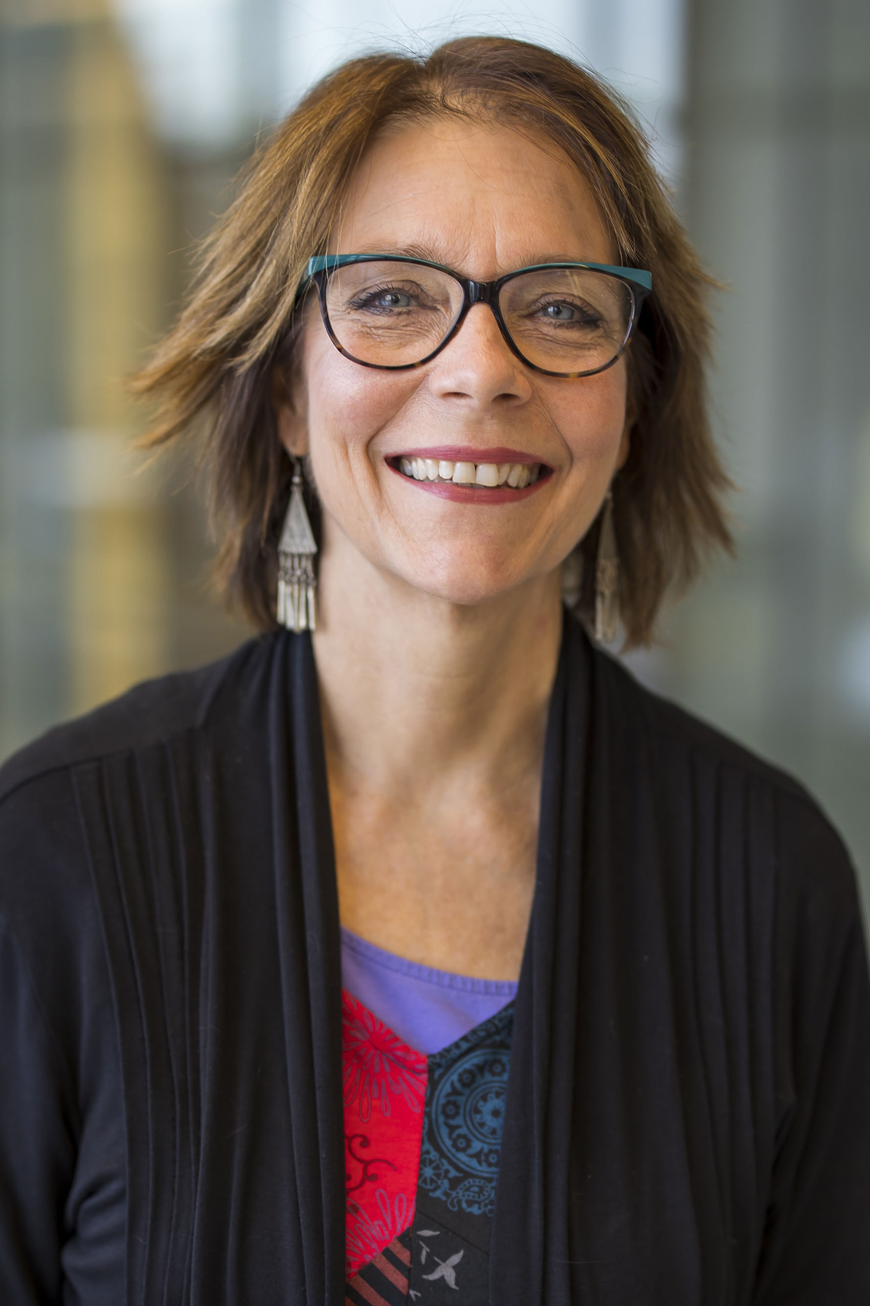 Callie Cary-Devine, a person with short brown hair wearing glasses, a red and blue shirt, and black cardigan, smiling while standing indoors with a blurred glass hallway in the background.
