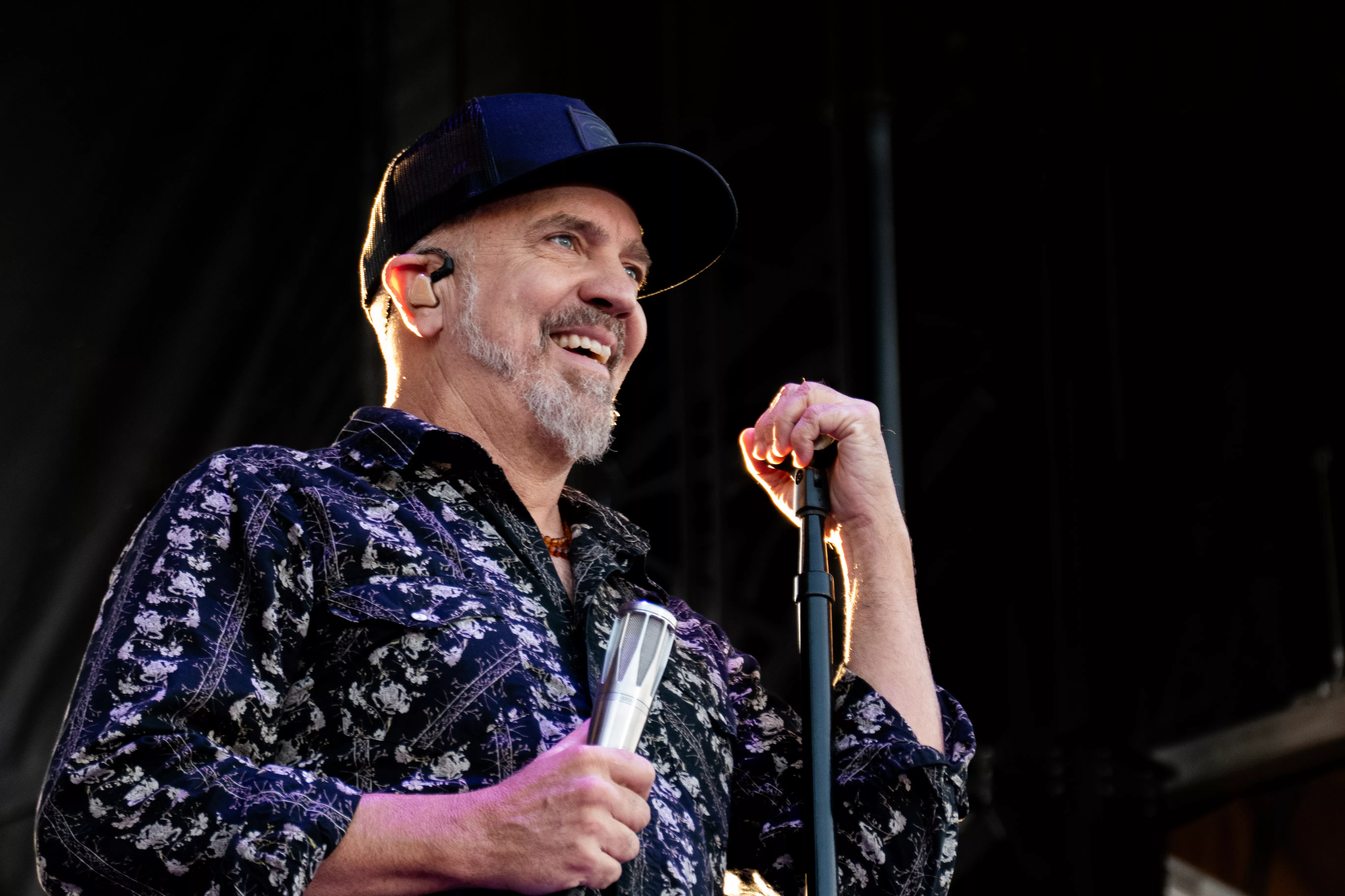 White male with grey beard, blue ballcap and blue print shirt holding a microphone