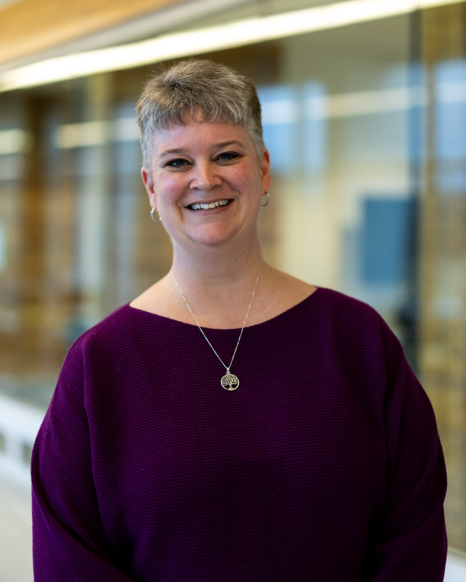 Erin Willoughby in purple long sleeve top with gold necklace smiling.