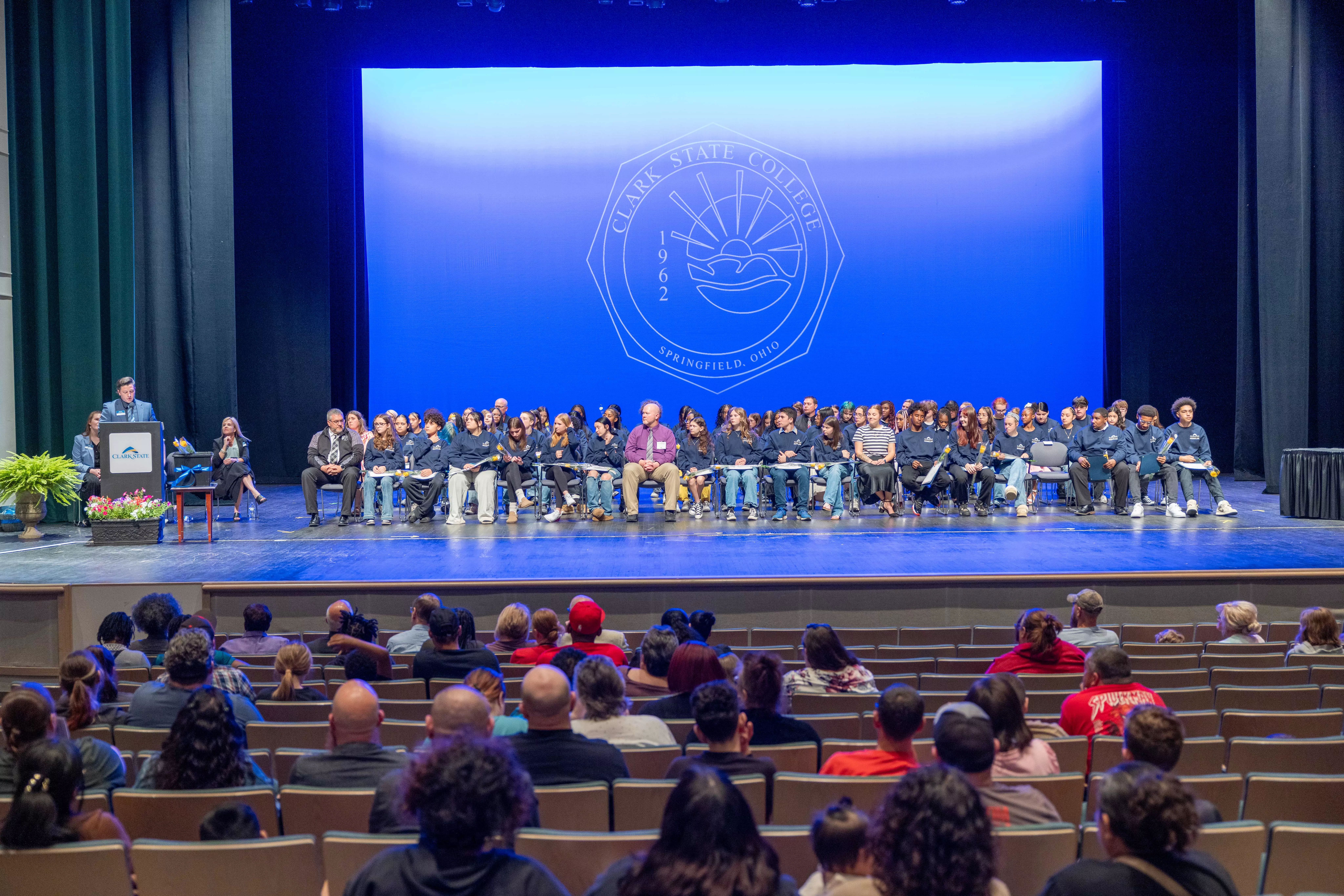 Stage filled with Scholars students, podium with audience in the foreground.