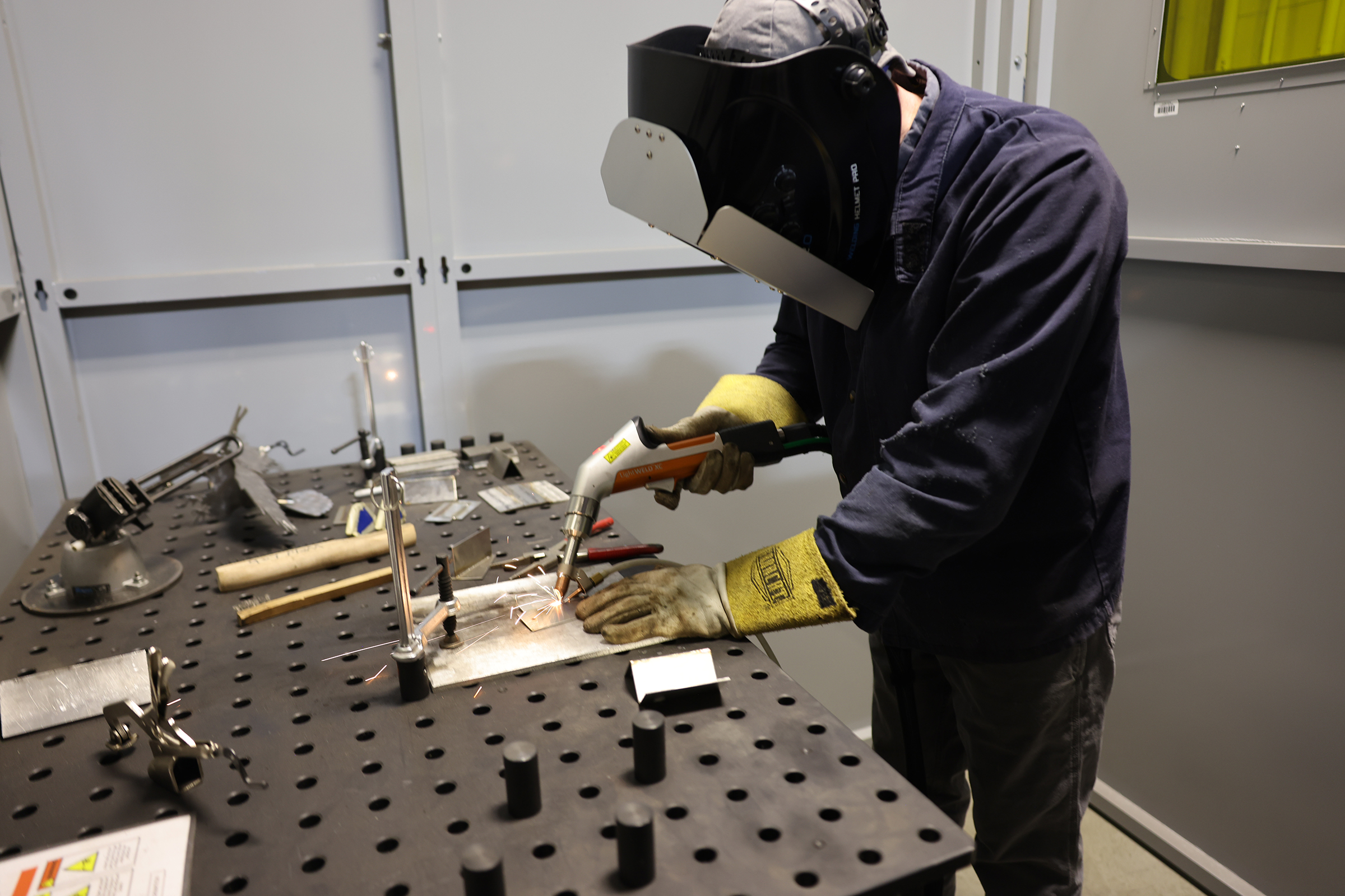 Student with welding helmet and yellow gloves uses handheld welding tool on workbench.