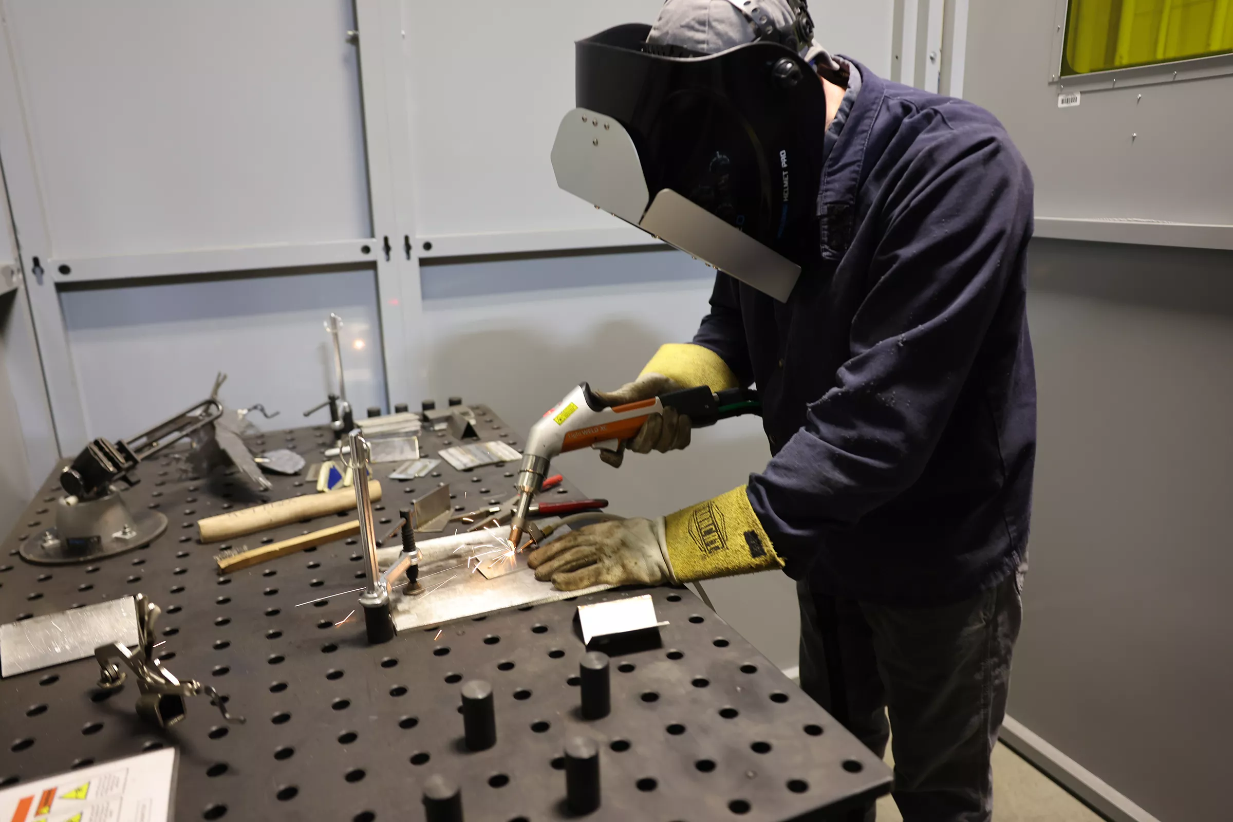 Student with welding helmet and yellow gloves uses handheld welding tool on workbench.