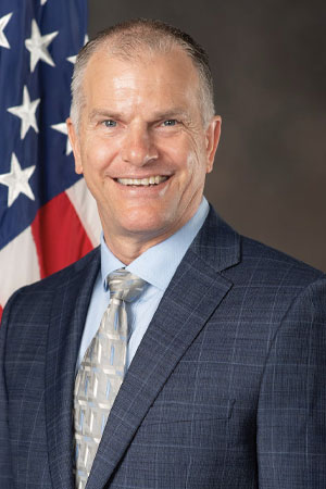 John Faulkner wearing a suite with an American flag behind him.