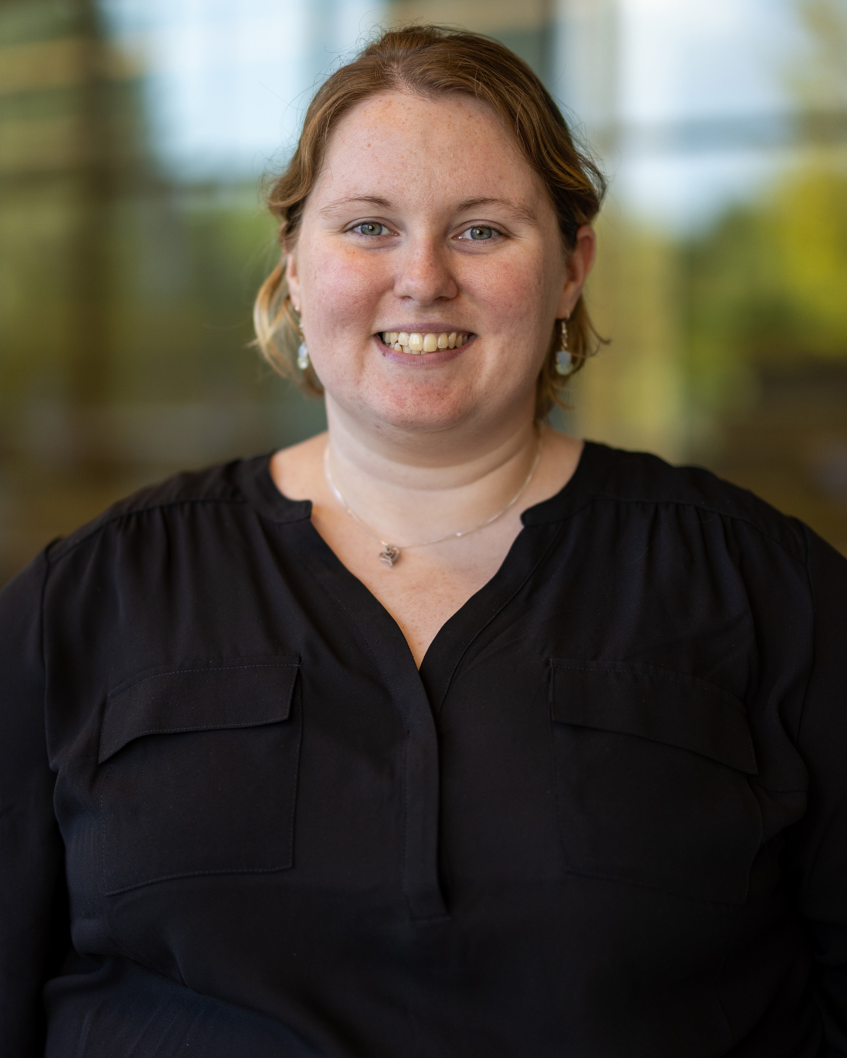 Whitney Deans, a person with short blonde hair wearing a black blouse, smiling while standing indoors with a blurred glass hallway in the background.